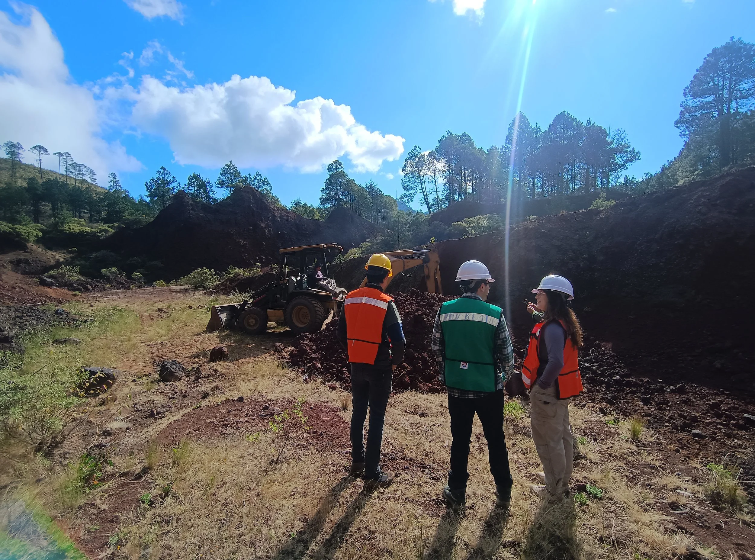 Tres personas con cascos y chalecos de seguridad observando maquinaria en una excavación en un paisaje de colinas y árboles.