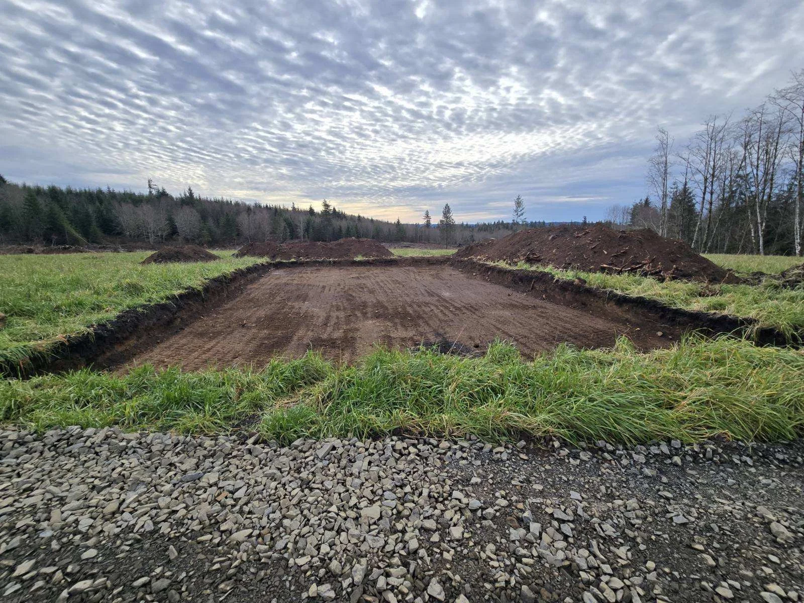 A construction site in a grassy field with a large open area of dirt, three piles of dirt at the back, surrounded by trees and a cloudy sky.