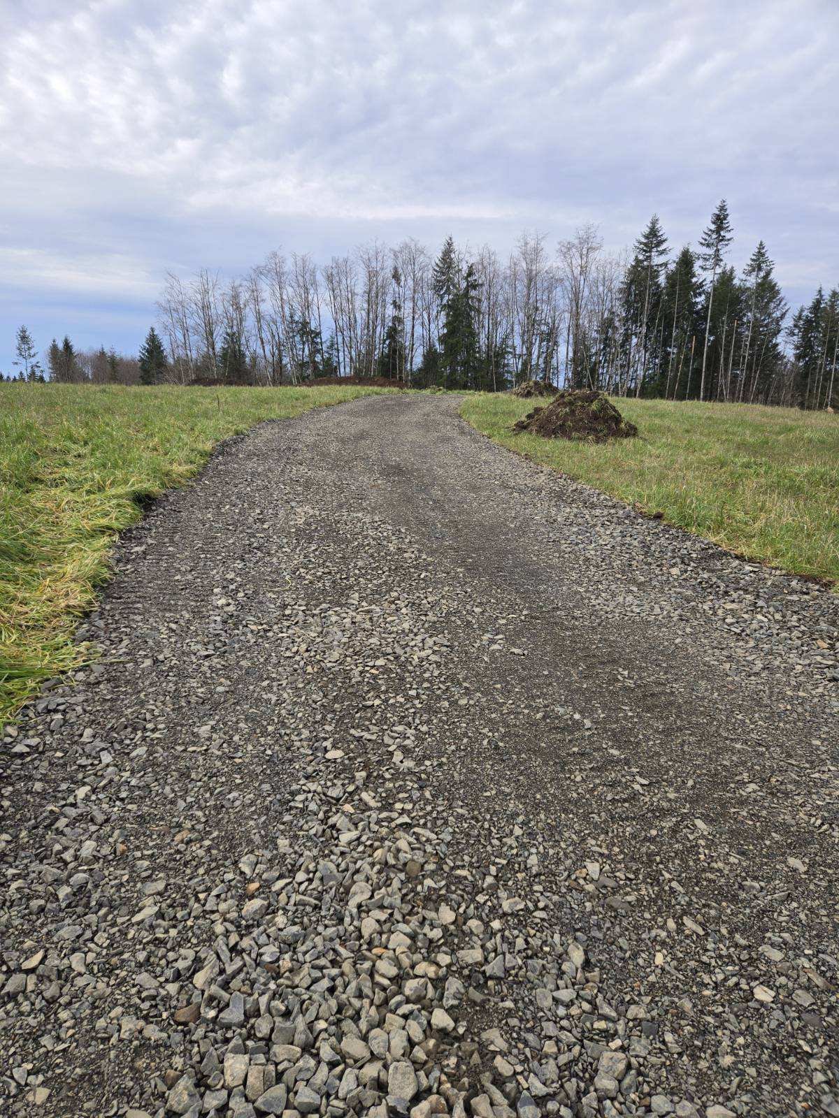 A gravel dirt road winding up a grassy hill with trees in the background under a partly cloudy sky.