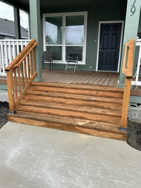 Front porch with wooden stairs and railing, two chairs, and a door with a window.