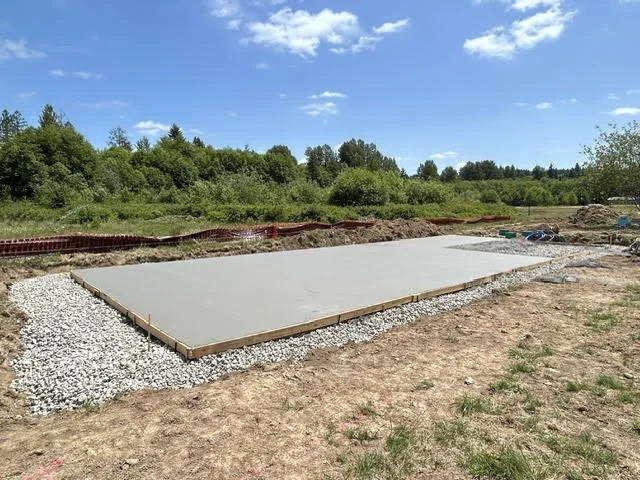 A freshly poured concrete slab foundation on a construction site surrounded by gravel and dirt, with a forested area and blue sky in the background.
