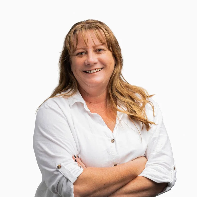 A woman with long, light brown hair smiling, wearing a white button-up shirt with rolled-up sleeves, standing with her arms crossed against a plain white background.