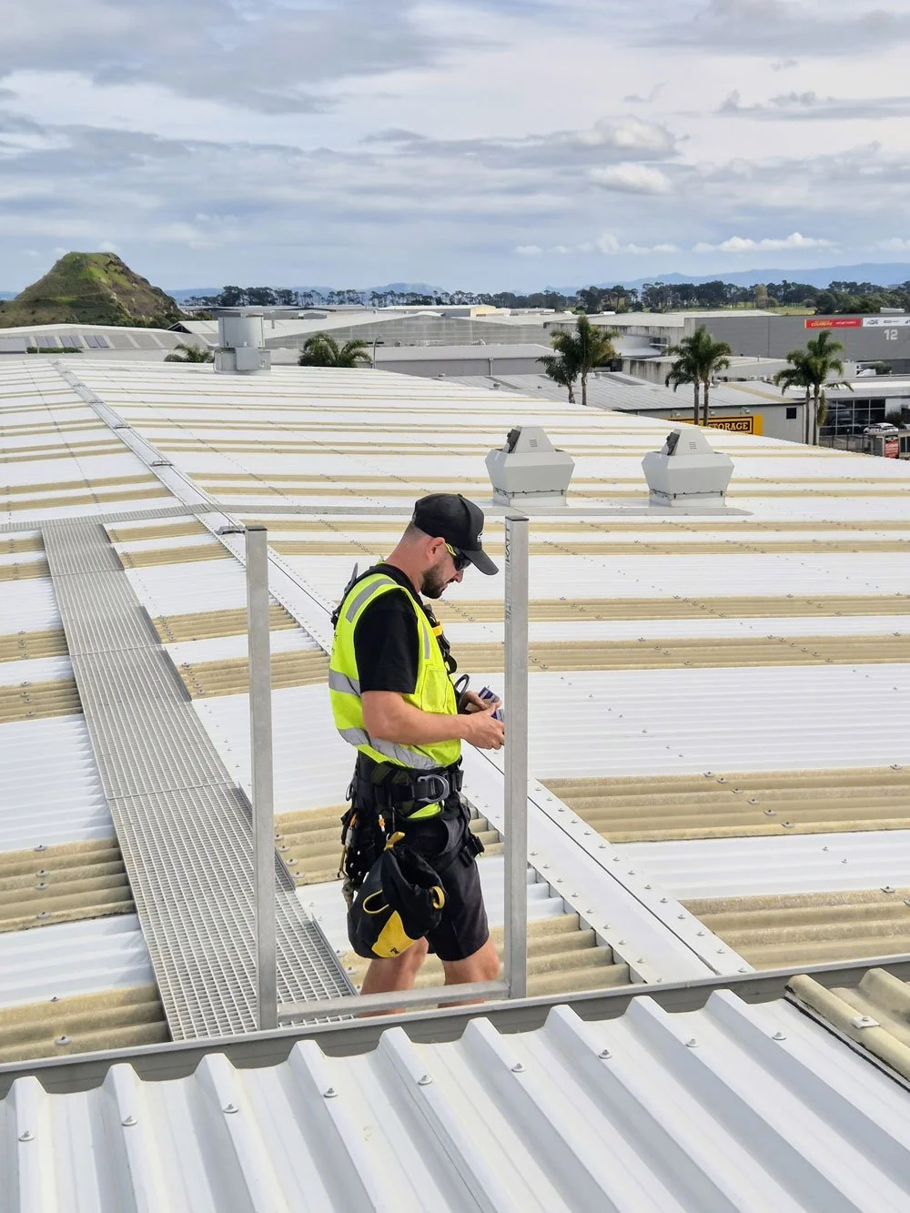 A worker in a safety vest and helmet standing on a rooftop, looking at a device in his hand, with a background of cloudy sky and distant industrial buildings.