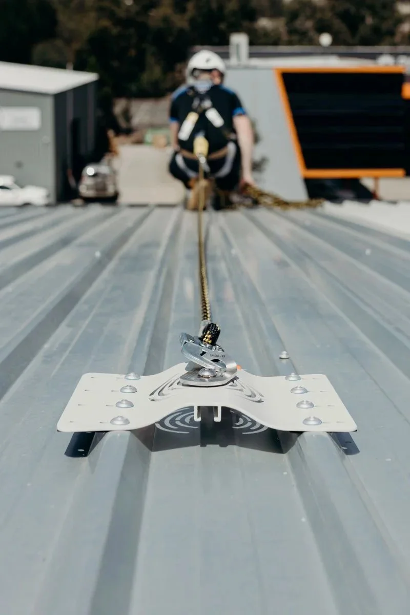 A person wearing safety gear, including a helmet and harness, is securing a rope to a rooftop on a metal surface using a Roof Anchor.