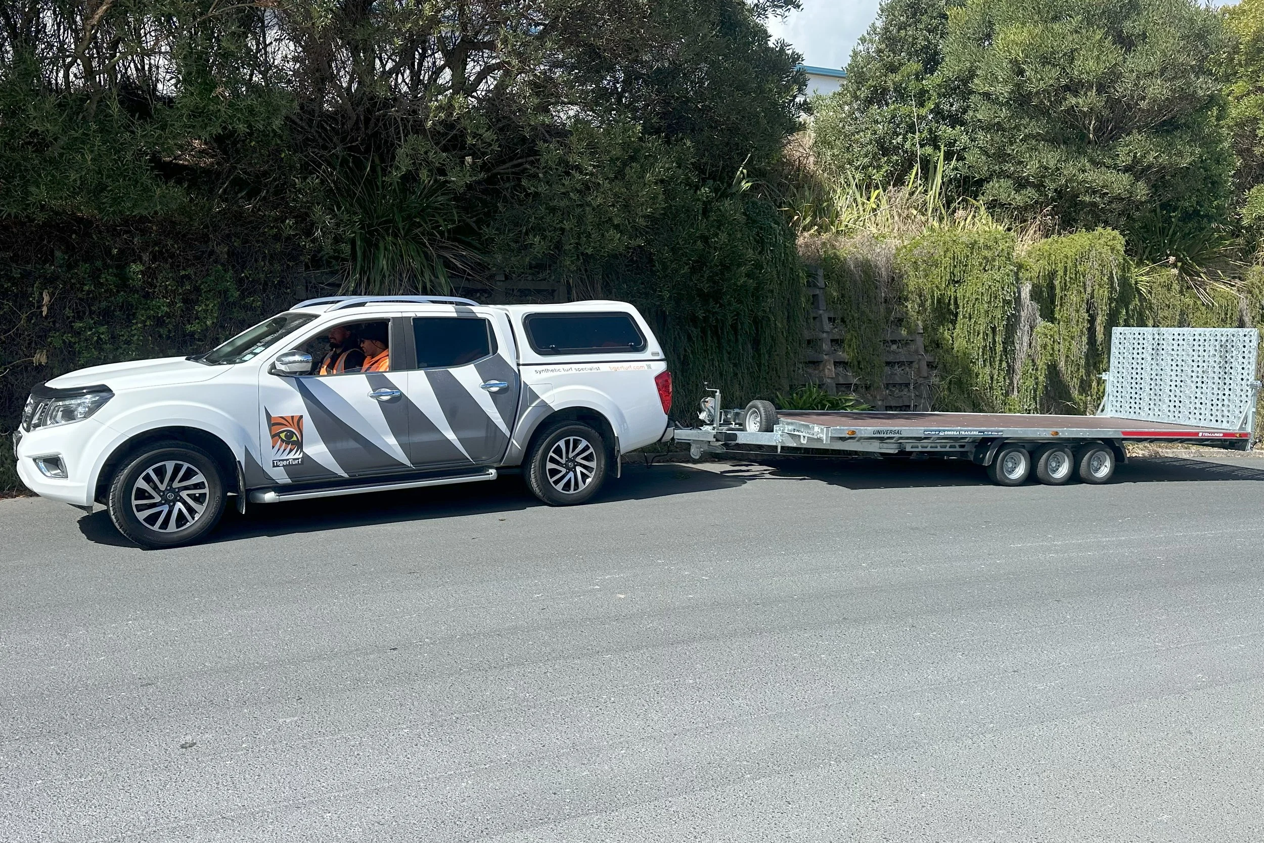 White pickup truck with graphics towing a flatbed trailer on a road beside greenery.