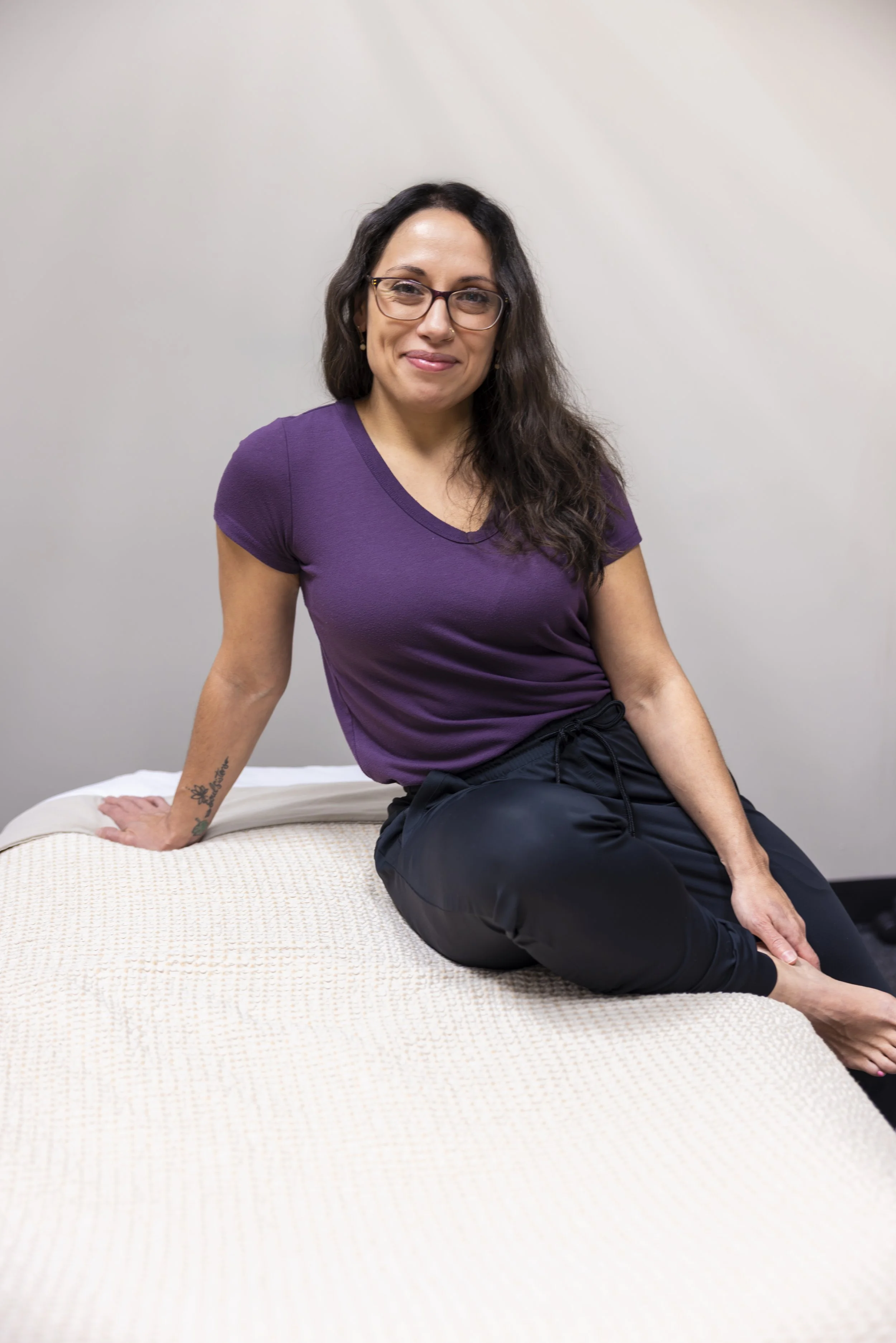Woman with glasses and long dark hair, sitting on a bed in a purple t-shirt and black pants, smiling at the camera.