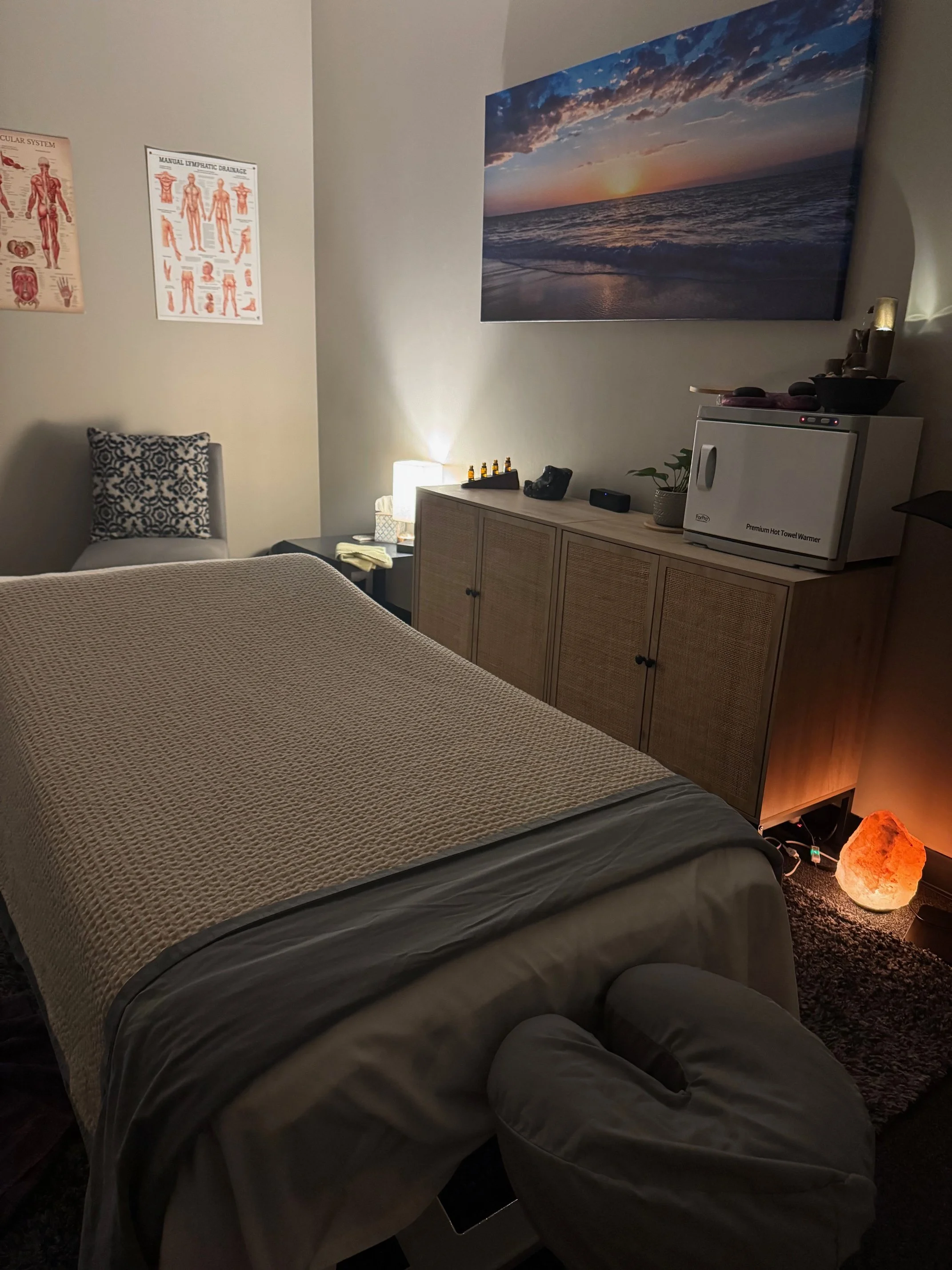 A massage room with a massage table covered in a beige blanket, a salt lamp on the floor, a large wall art of a beach sunset, a cabinet with a salt warmer, essential oils, and a small plant, and a chair with patterned cushion in the corner. posters o