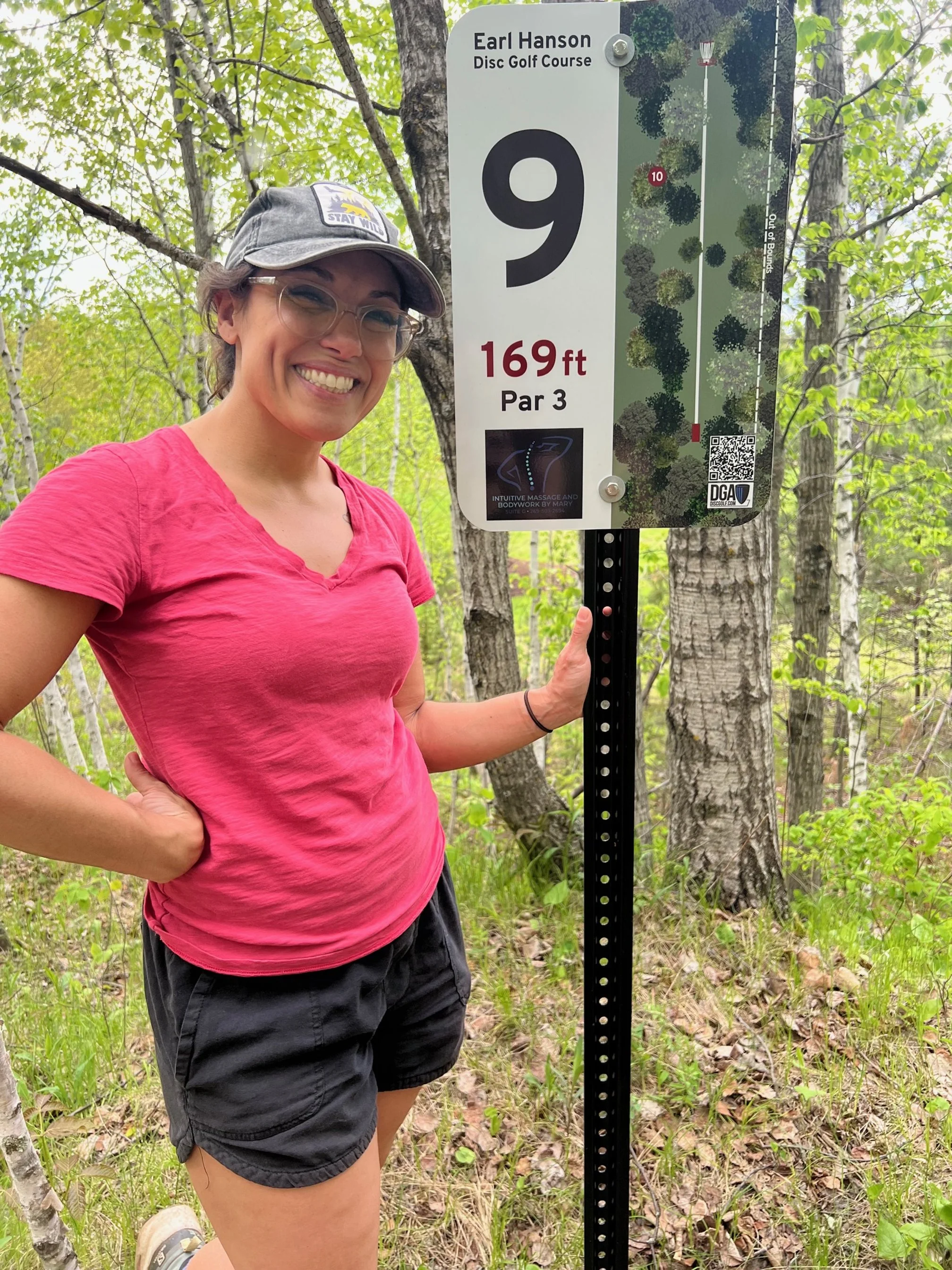 A smiling woman with glasses, a gray cap, and a pink shirt standing outdoors next to a disc golf course sign that shows hole 9, 169 feet, par 3, located in a wooded area with trees and greenery.