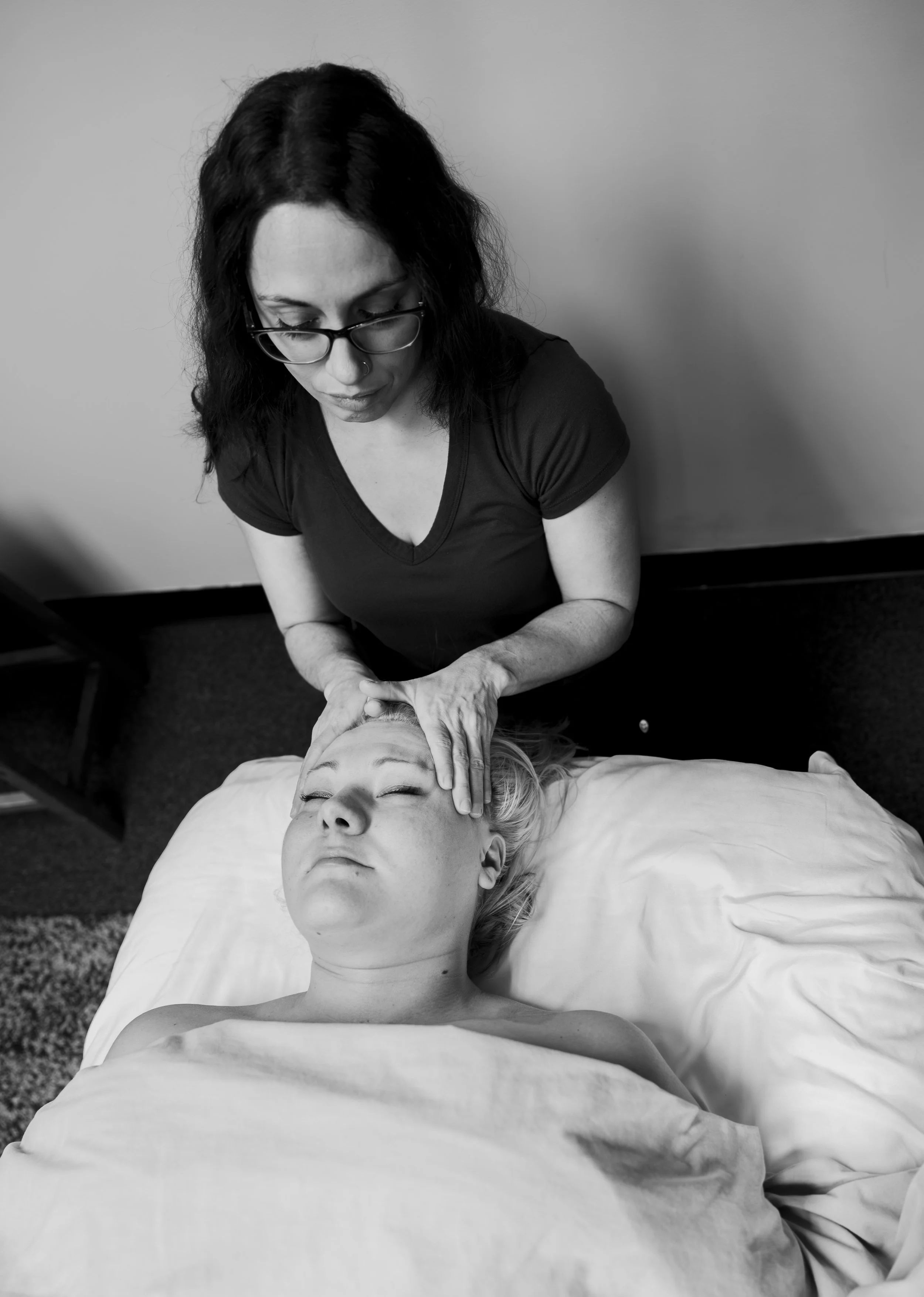 Black and white photo of a woman giving a facial massage to a young woman lying in bed with her eyes closed.