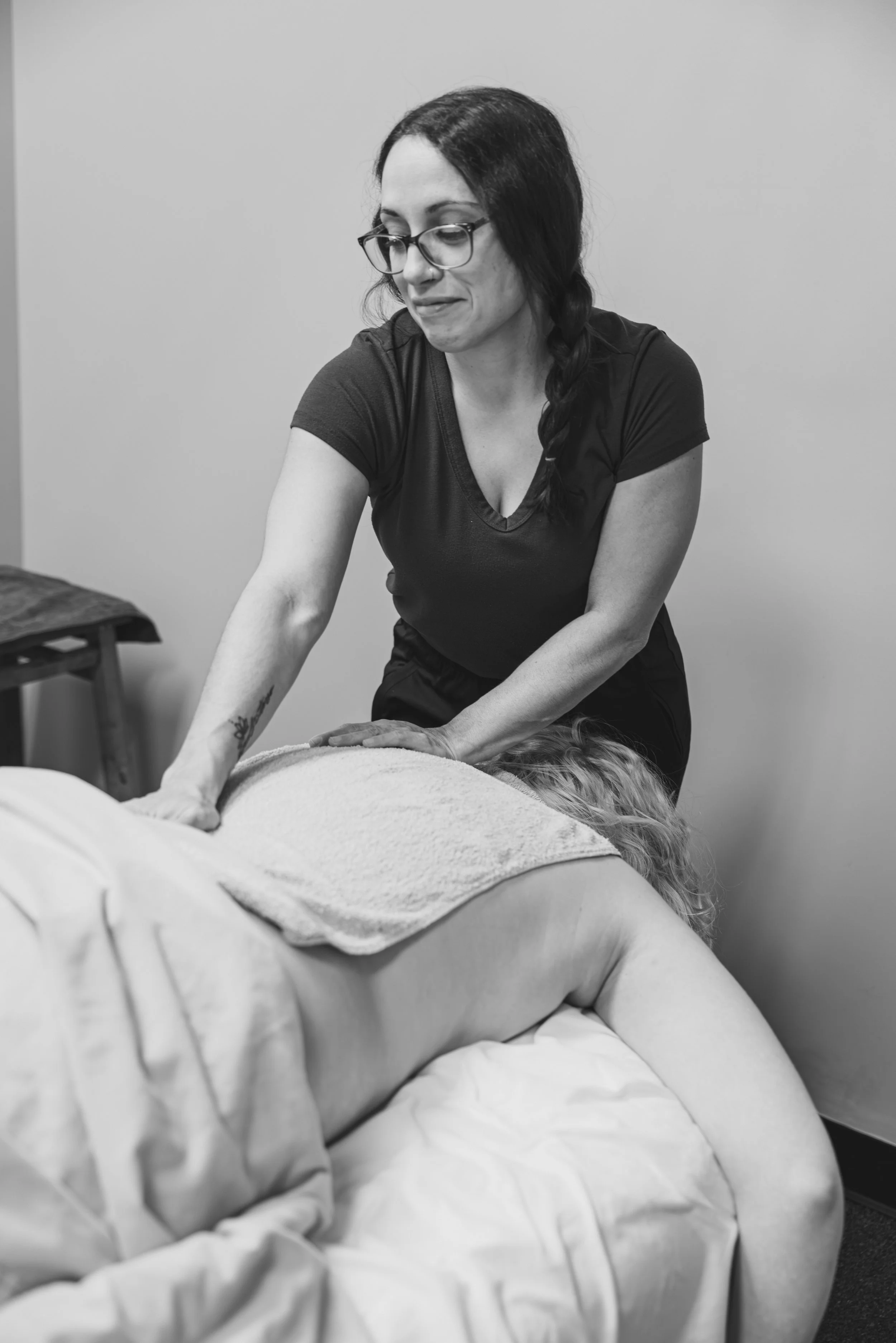 A woman giving a massage to a person lying face down on a bed, with a towel on their back, in a massage room.