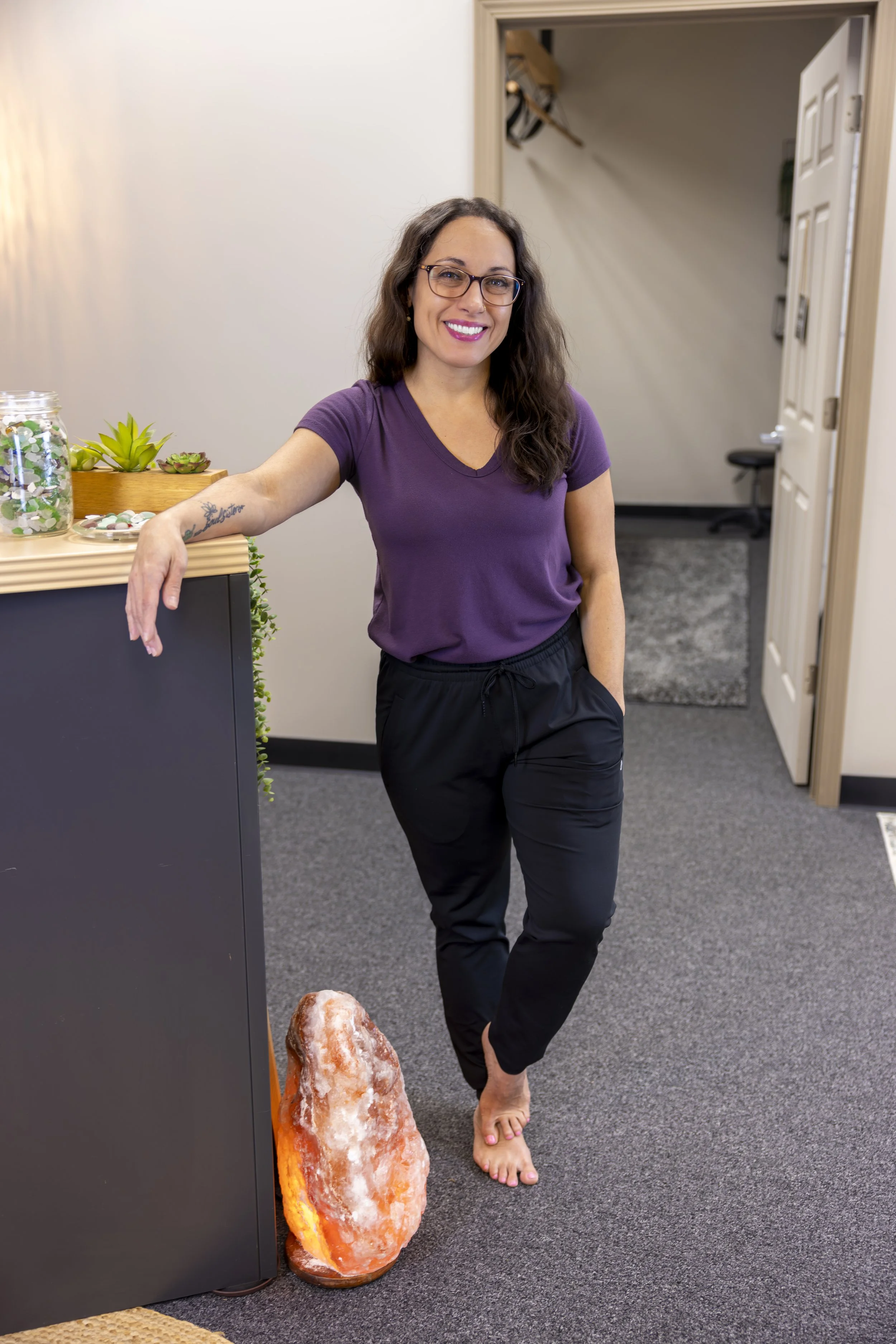 A woman with glasses and long brown hair, wearing a purple t-shirt and black pants, standing barefoot in an office. She is smiling and leaning against a gray counter with her arm resting on it. There are small plants and decorative rocks on the count