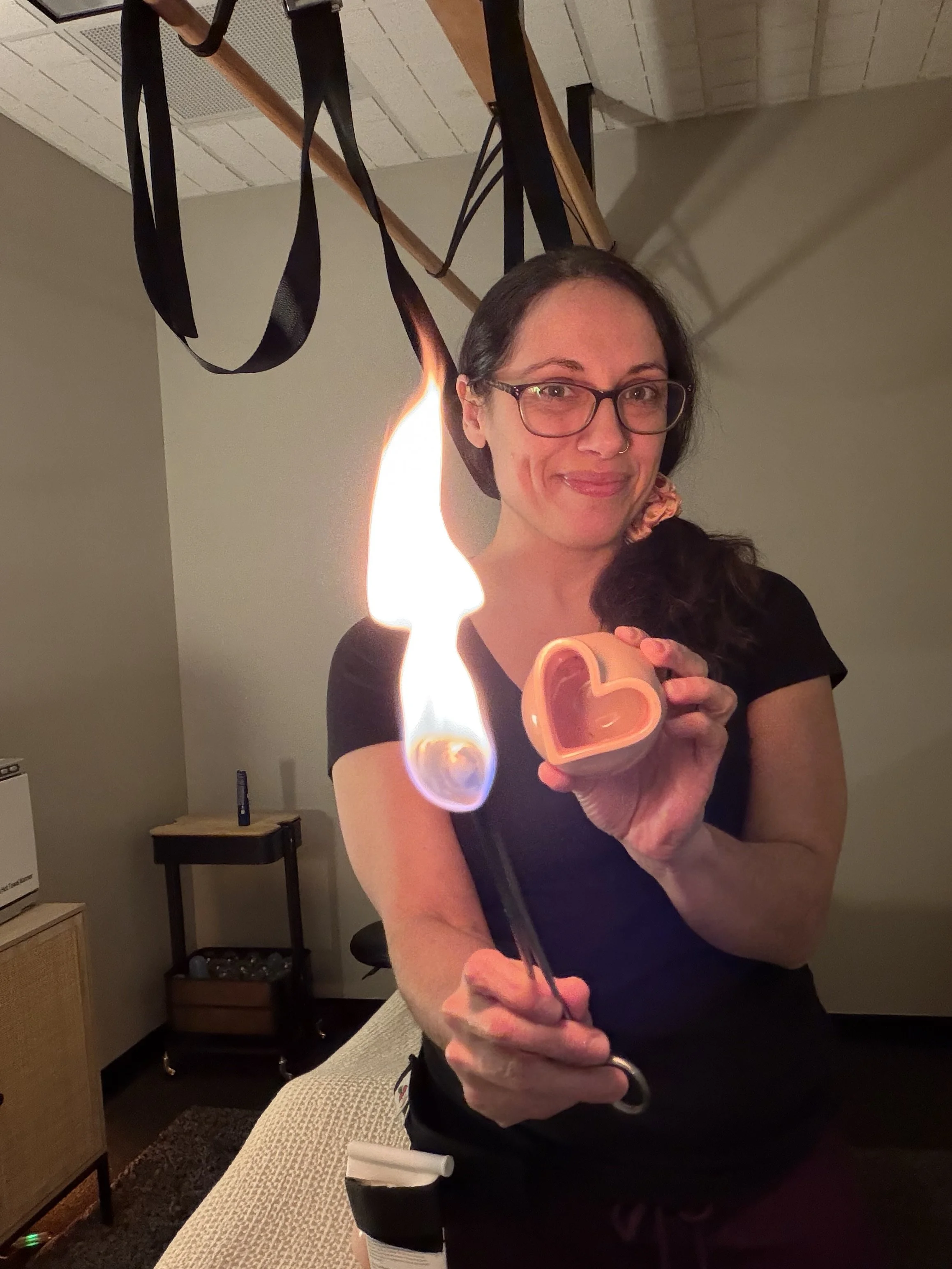 Woman with glasses and a nose ring holding a heart-shaped lighter with a flame, in a room with beige walls and furniture.