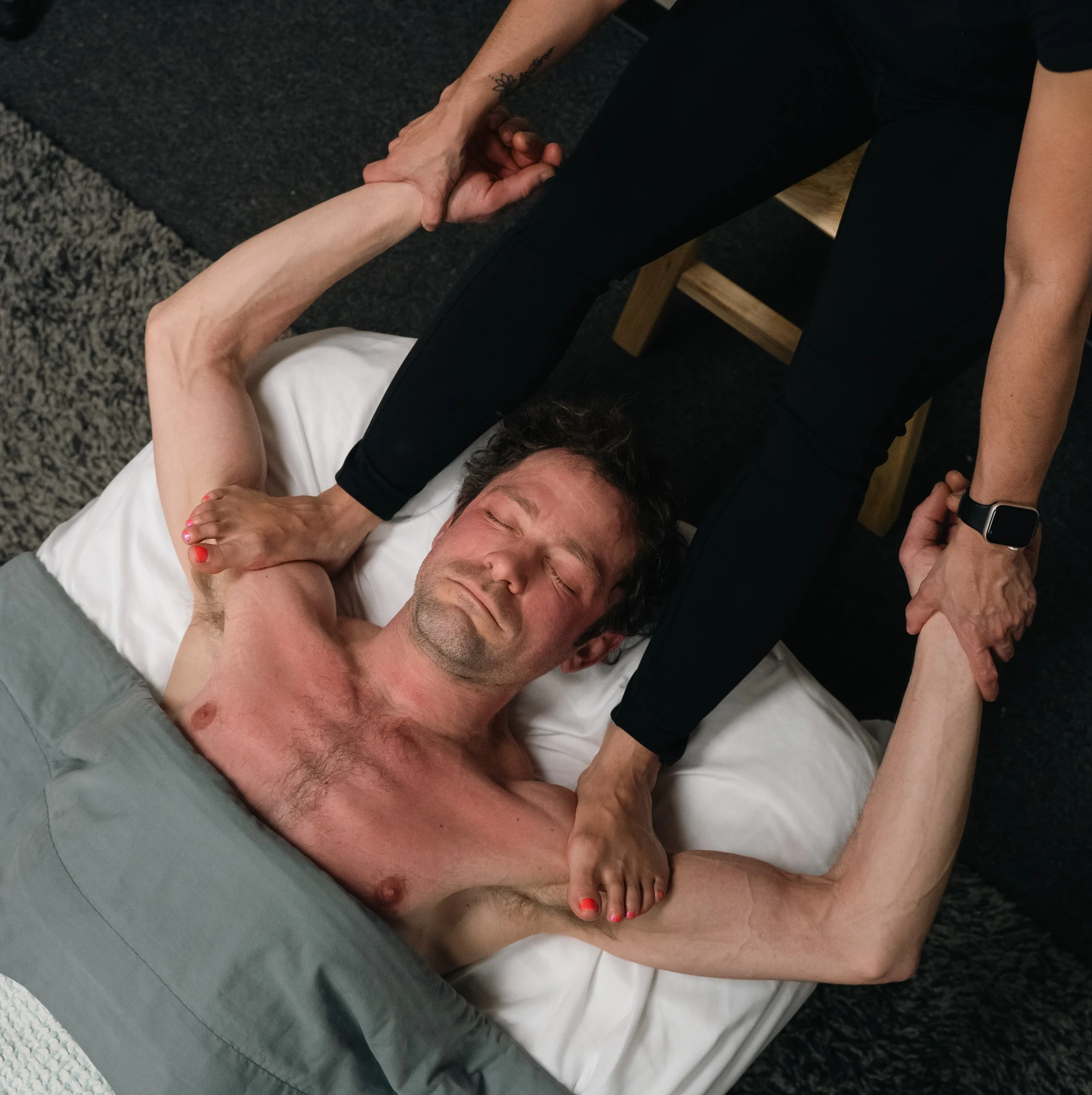 A man lying shirtless on a bed with eyes closed, holding hands with a woman who is standing and leaning over him giving barefoot massage on shoulders.