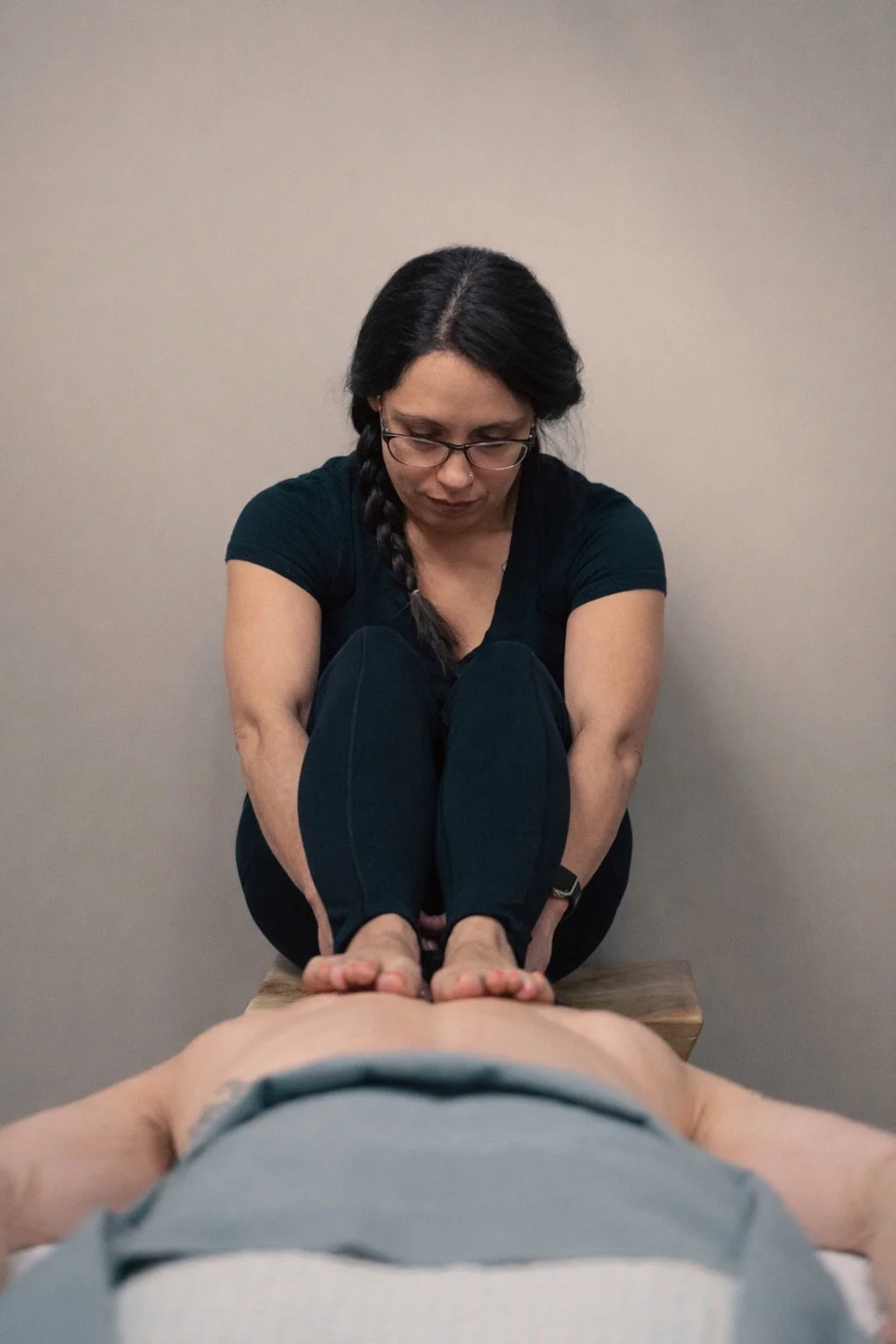 Woman with glasses receives a physical therapy or massage, sitting on a stool with a bare back, while a therapist applies pressure with her hands on the woman's upper back and shoulders.