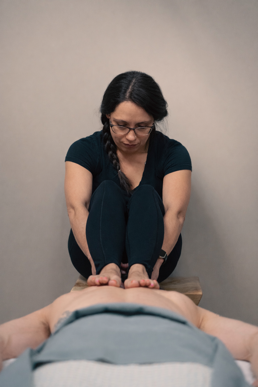 A woman with glasses and dark hair braided over her shoulder is performing a barefoot massage on a person's back.