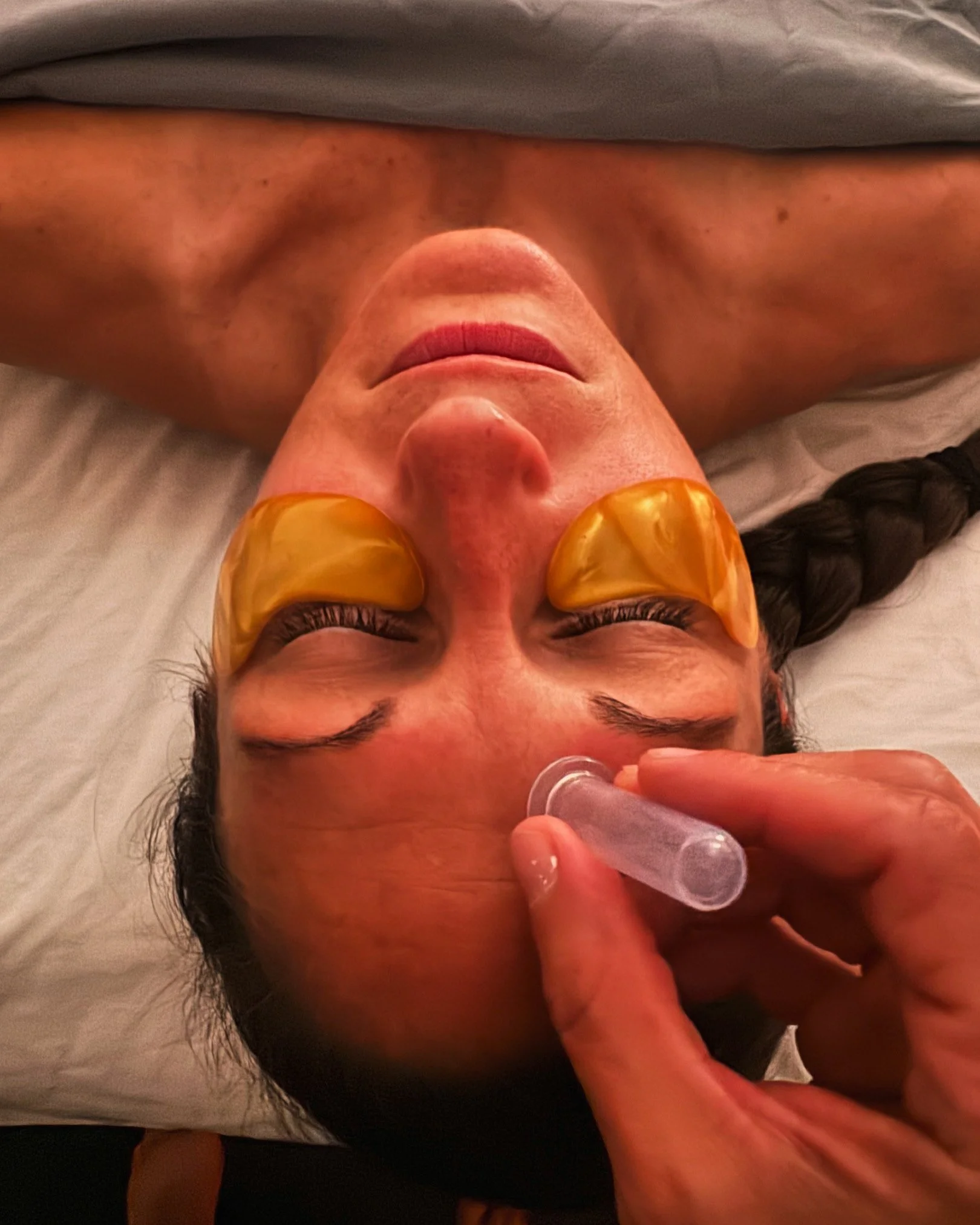 Woman lying on a bed with her eyes closed, receiving a facial treatment with gold eye masks and an ultrasonic facial device