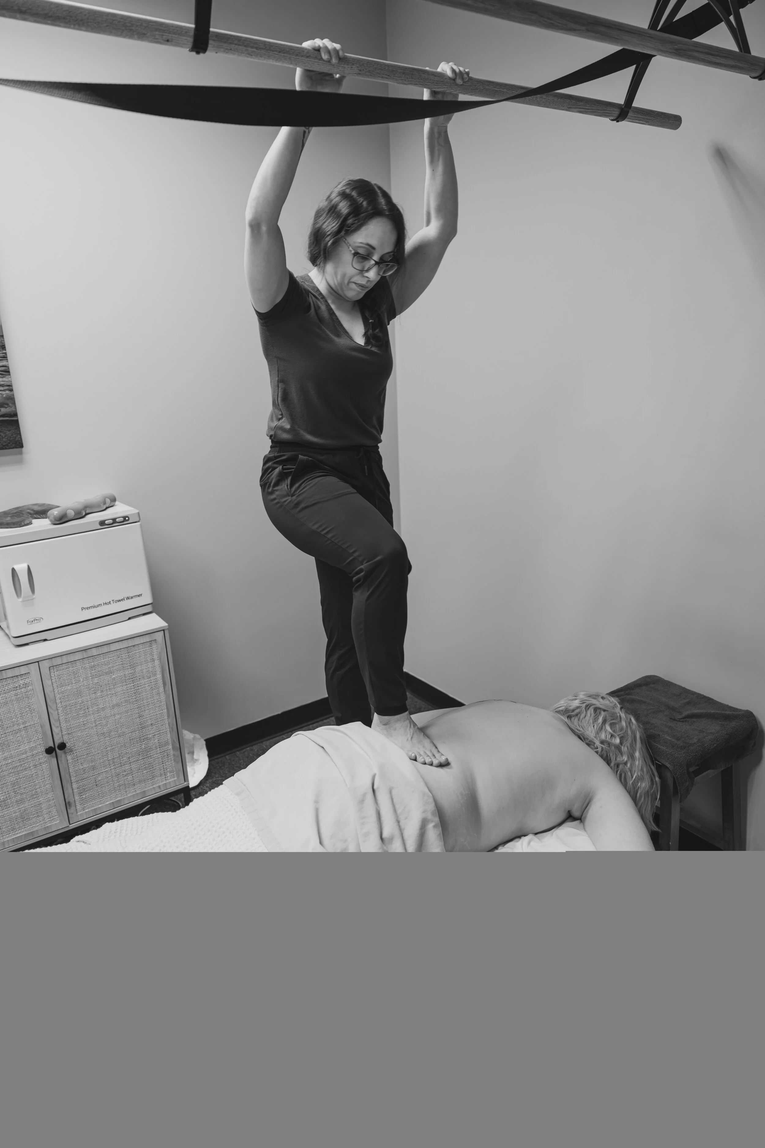 A massage professional is performing barefoot massage on a client's back during a therapy session or massage, in a room with a cabinet and a hot towel warmer.
