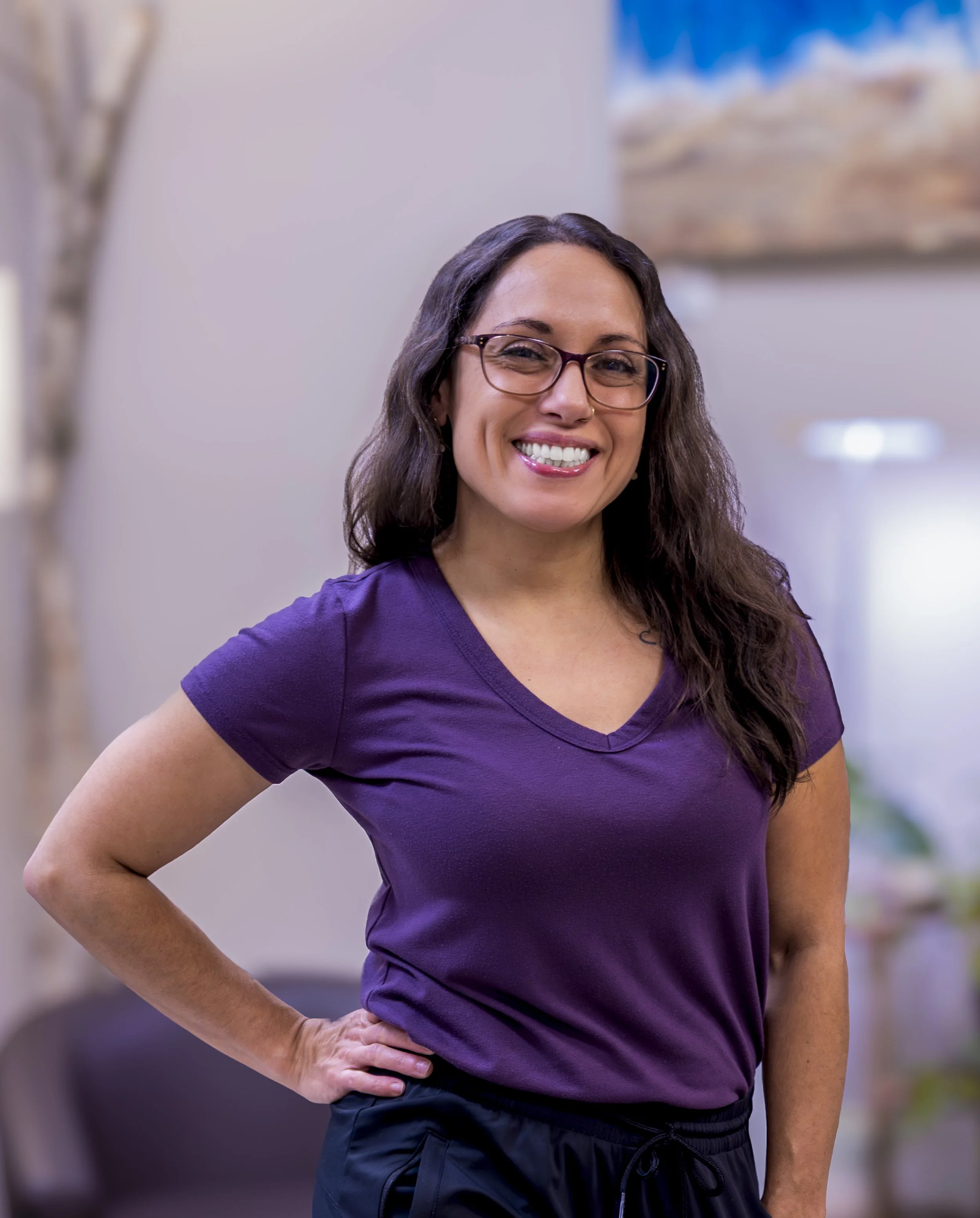A woman with glasses and long dark hair, smiling, wearing a purple short-sleeved shirt and black sports shorts, standing indoors with one hand on her hip.