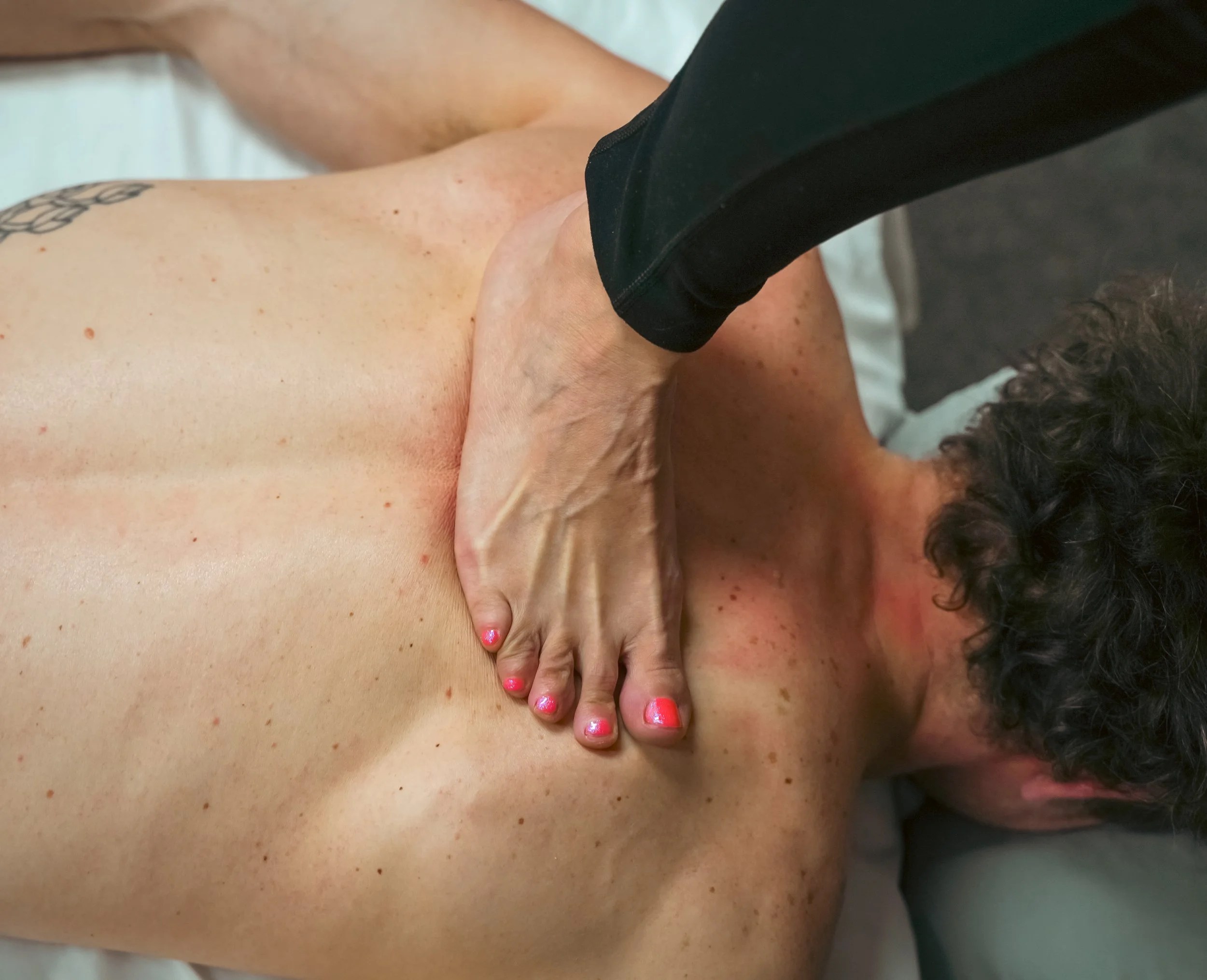 A person receiving a barefoot massage  on their upper back and neck area, laying face down with a therapist applying broad, consistent pressure with their feet.