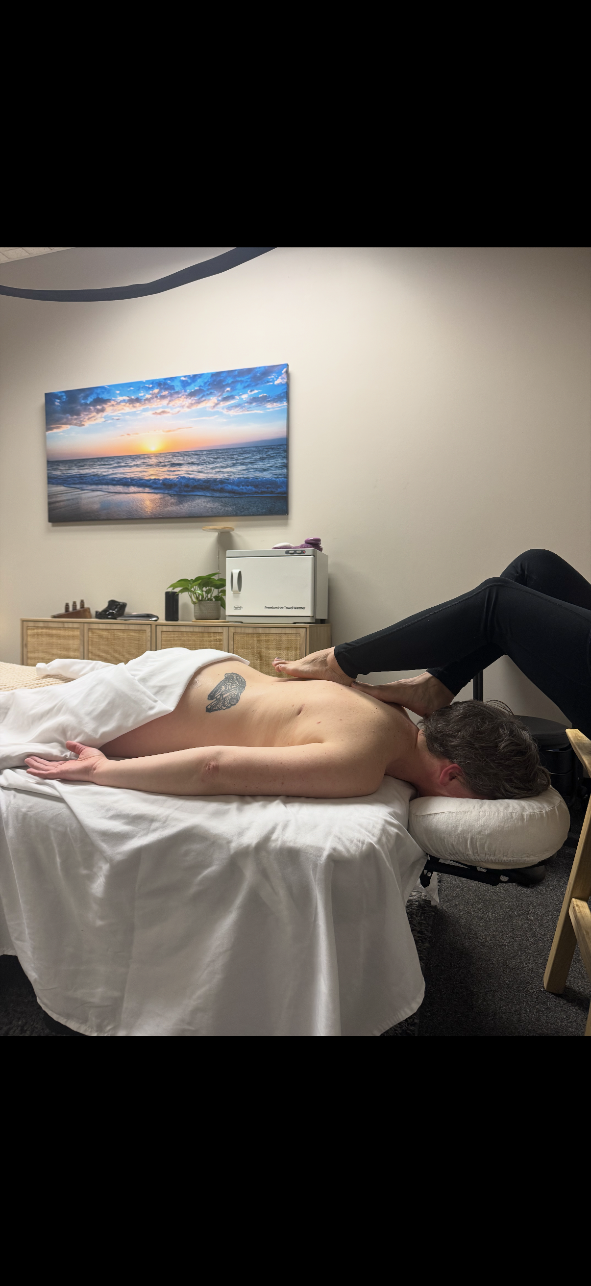 A person lying face down on a massage table receiving a barefoot massage in a room with a beach sunset picture on the wall and a small cabinet with plants in a quiet, cozy spa setting.