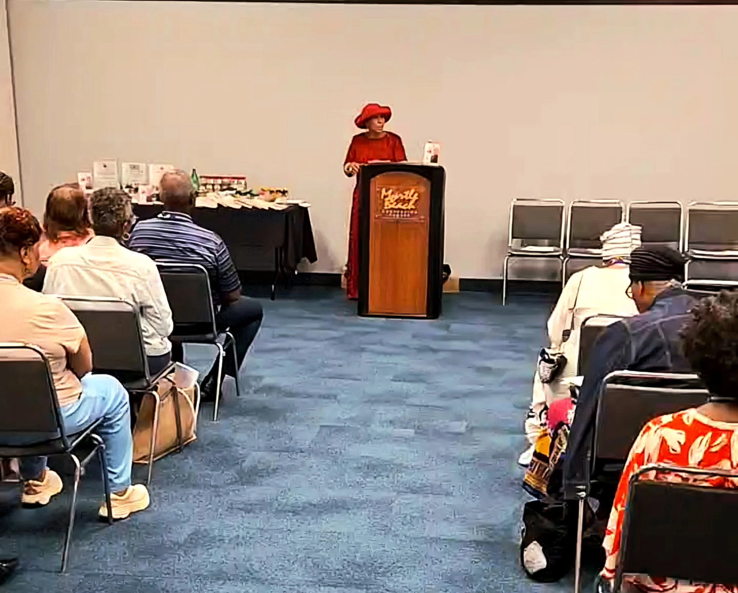 Flower arrangements with pink, white, and peach roses on metal stands decorate an outdoor wedding aisle lined with wooden chairs, leading to a white draped archway with greenery in the background.