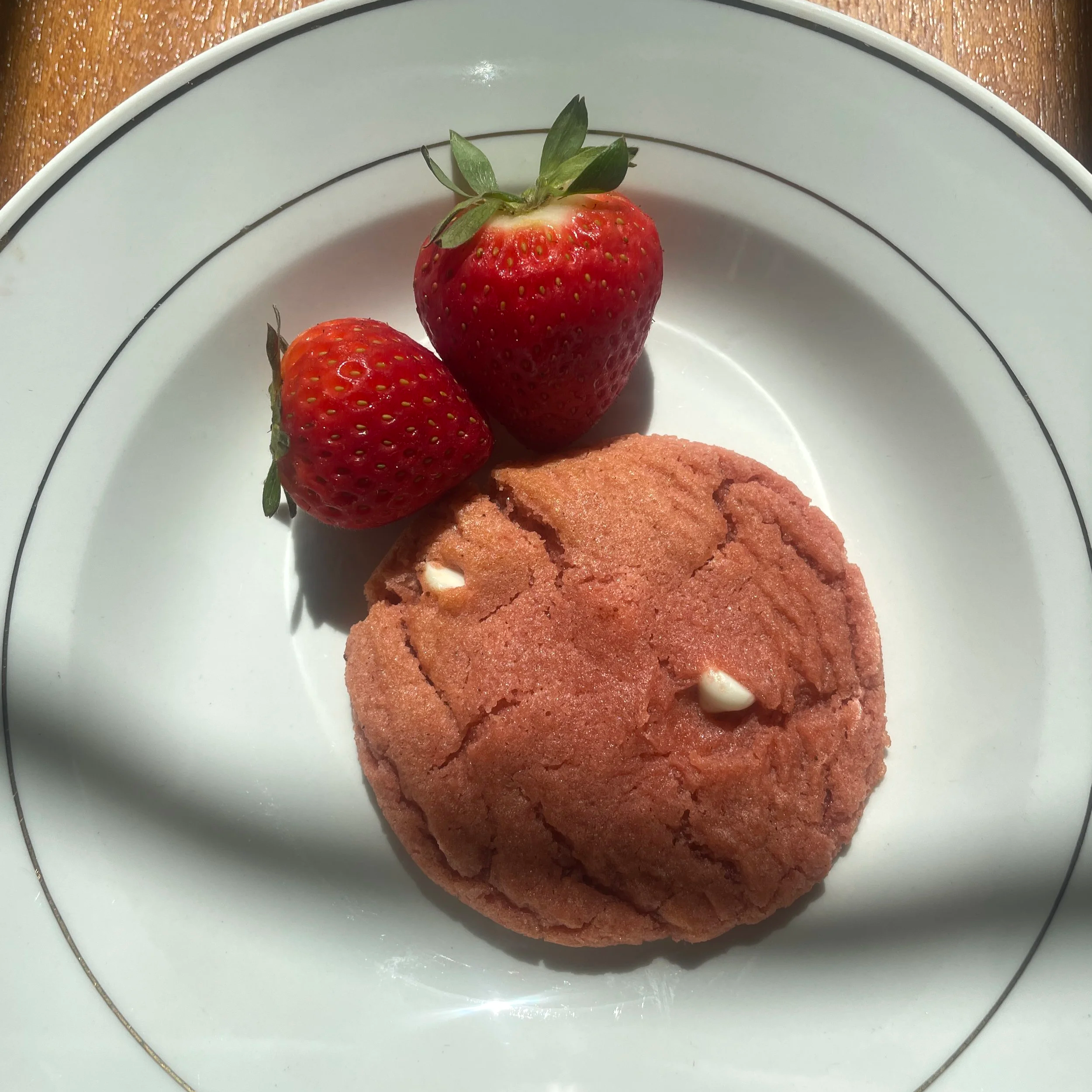 A white plate with two strawberries and a pink cookie with white chocolate chips on a wooden table.