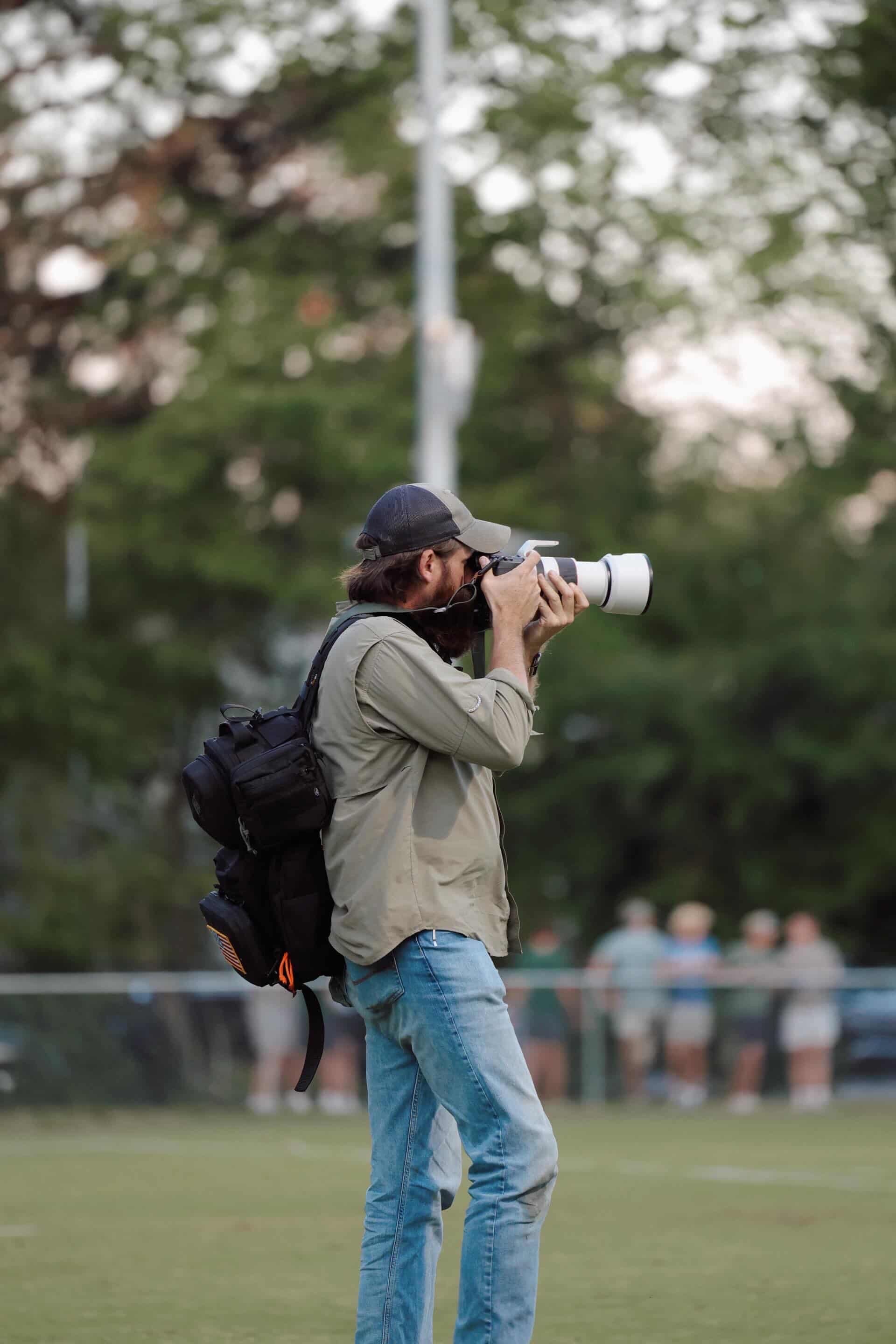 A man with long hair, glasses, gray jacket, jeans, and a black backpack takes a photo with a white camera outdoors with a blurred background of trees and a group of people.