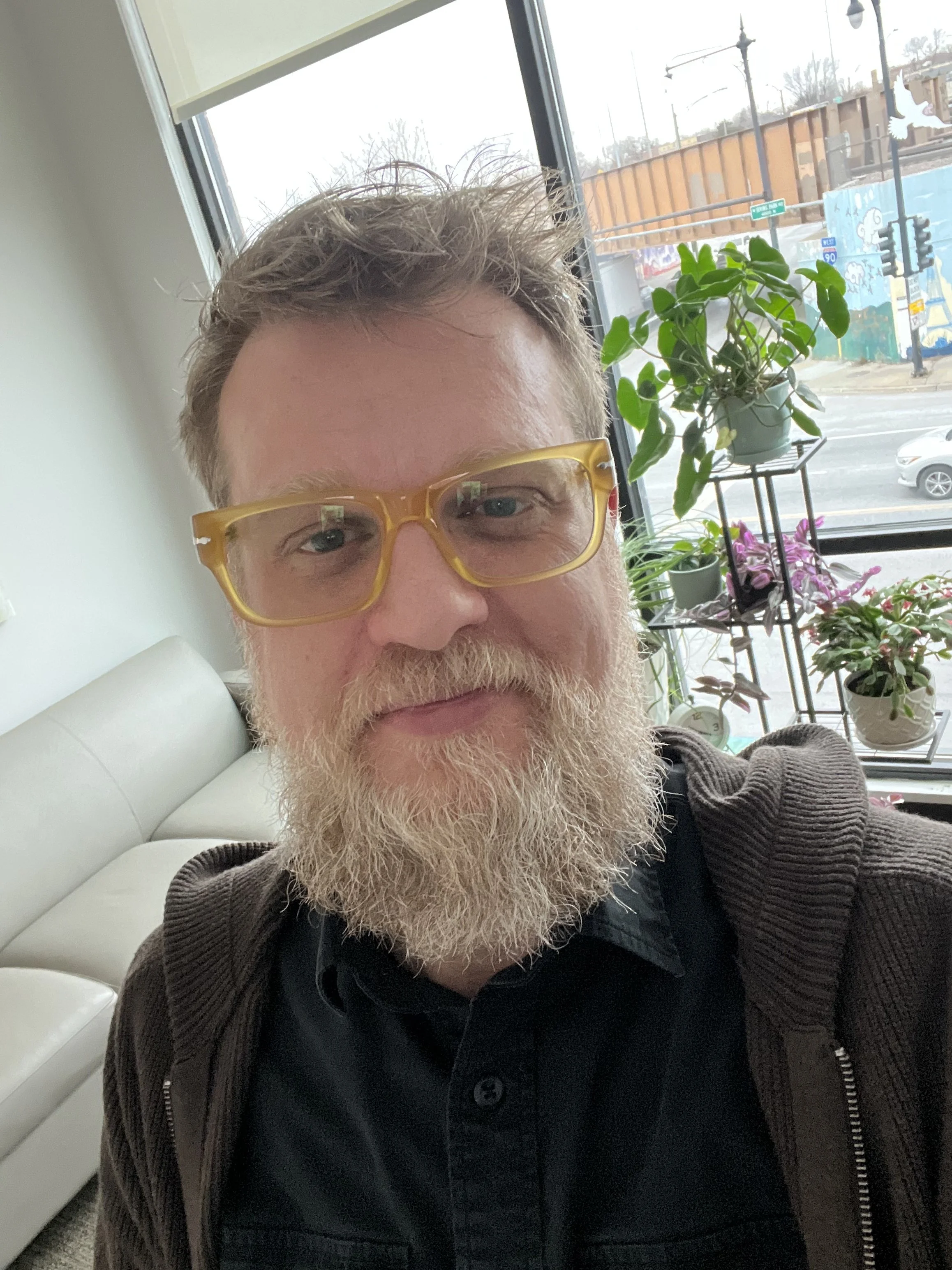 Close-up of a middle-aged man with gray hair, a beard, and glasses with yellow frames, sitting indoors near a window with plants outside.