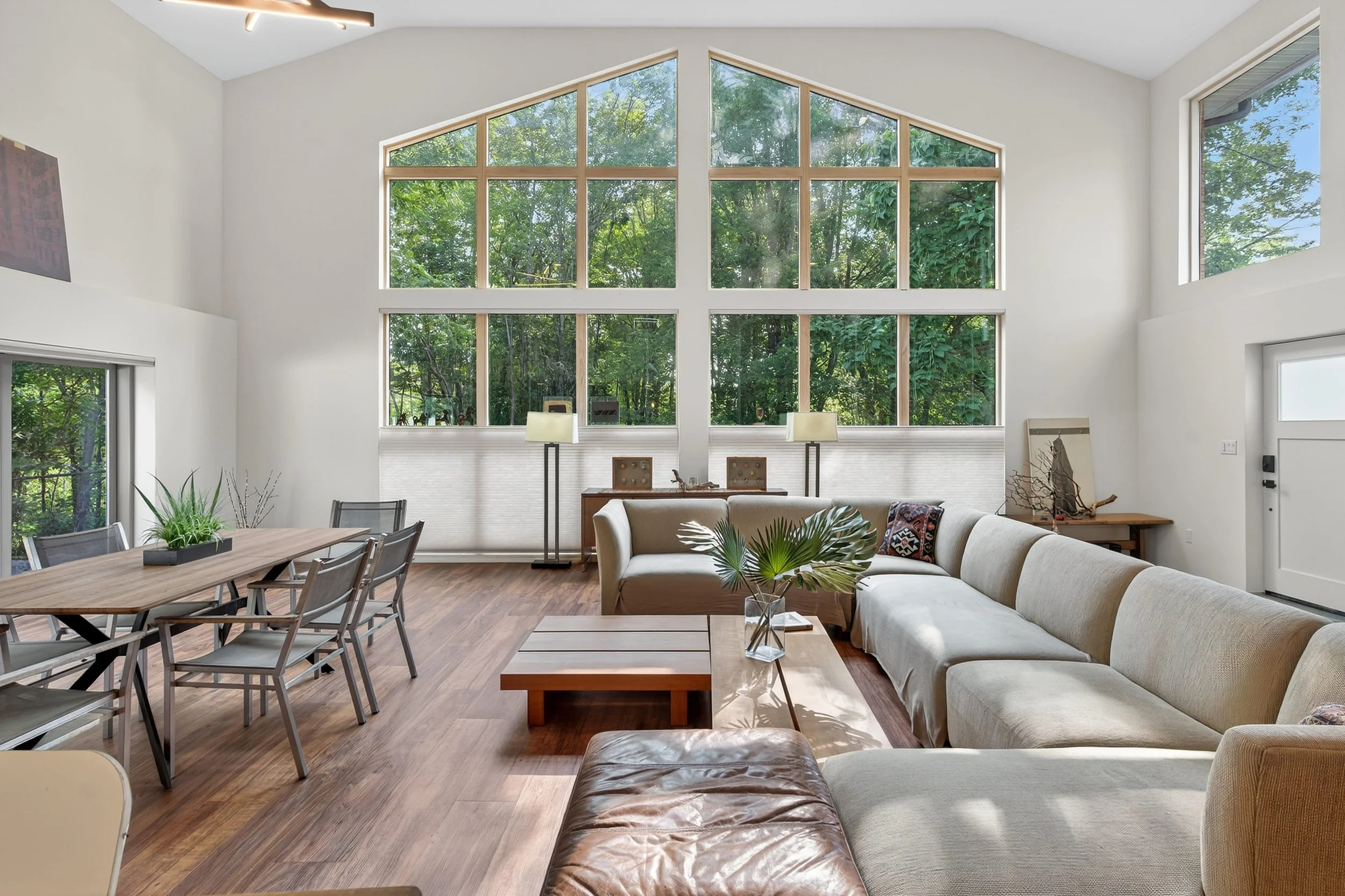 Interior view of a modern living room with large triangular window overlooking green trees, featuring a beige sectional sofa, wooden coffee table, and dining area with a wooden table and metal chairs.
