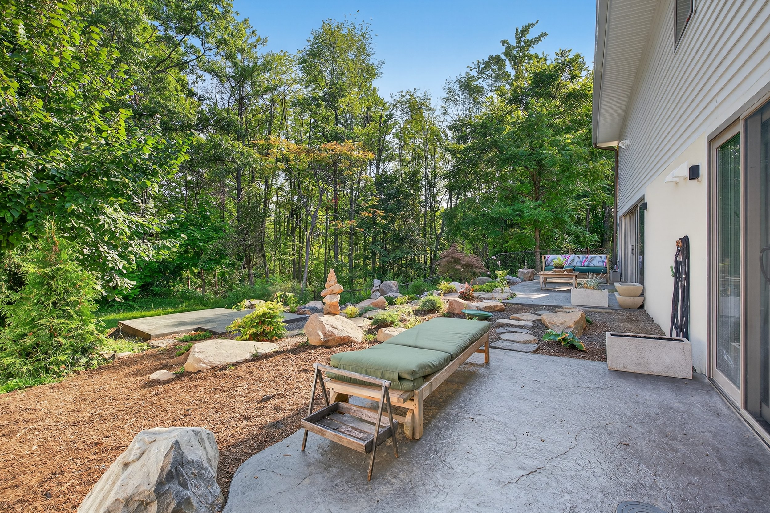 A backyard patio with outdoor lounge chairs, a garden with rocks, trees, and a house with large windows.