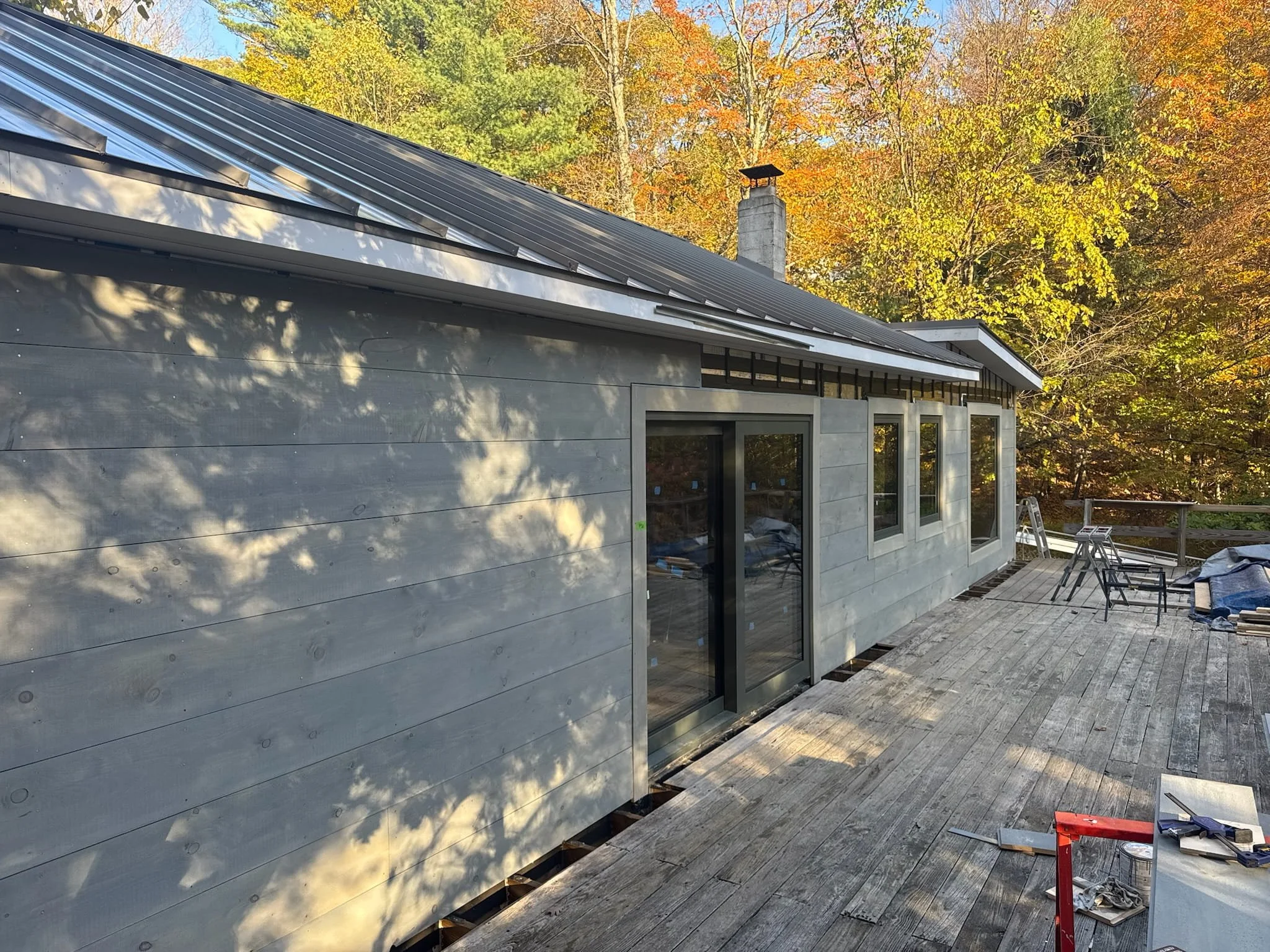 Partially constructed house with a sloped metal roof, large windows, and a wooden deck in an outdoor setting with trees showing fall foliage.
