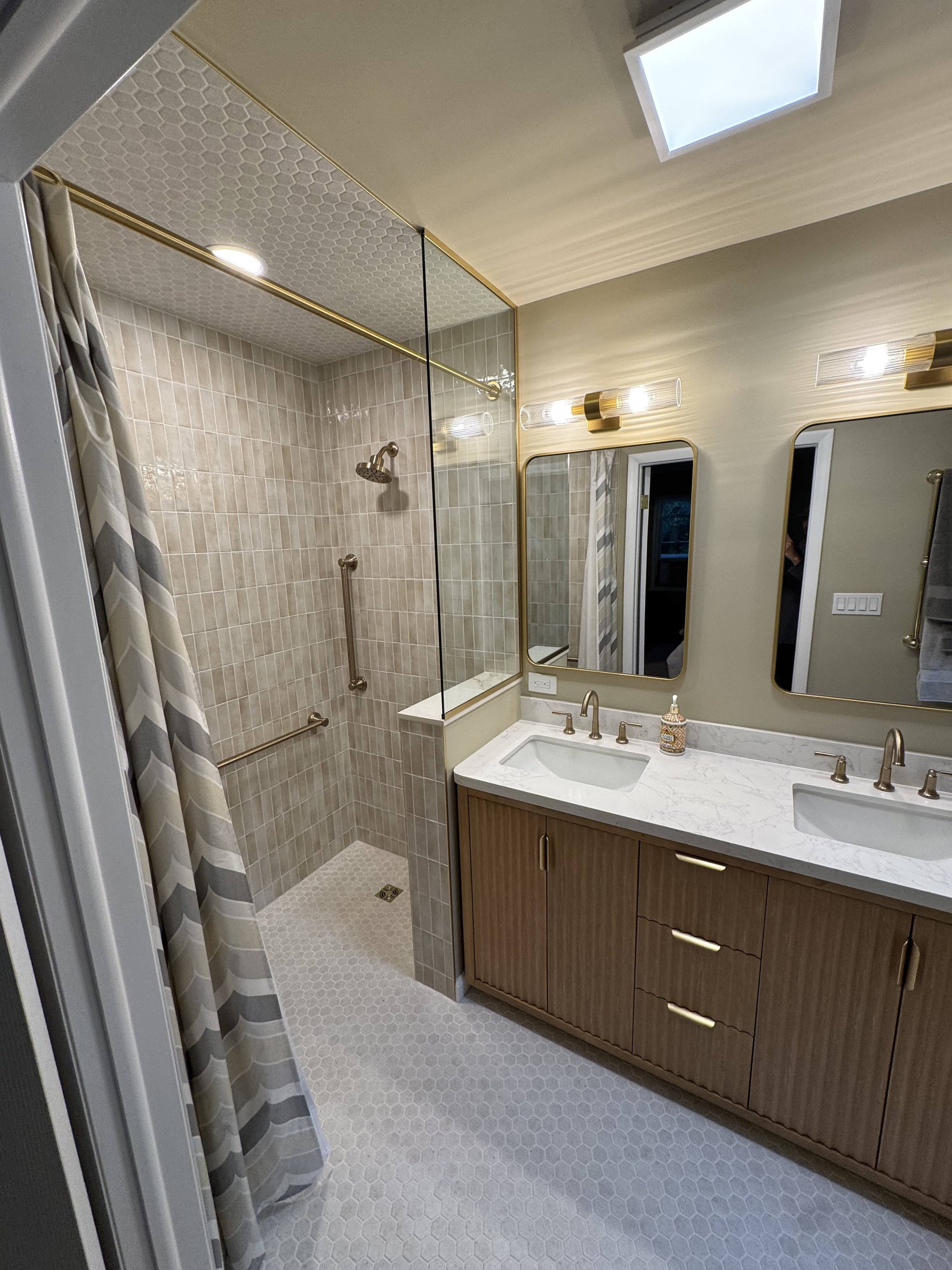 A modern bathroom with a shower area that has a glass partition, beige tiles, and a rain showerhead. A wooden vanity with a white marble countertop and double sinks, large mirrors with gold frames, and contemporary light fixtures. Skylight and window add natural light.