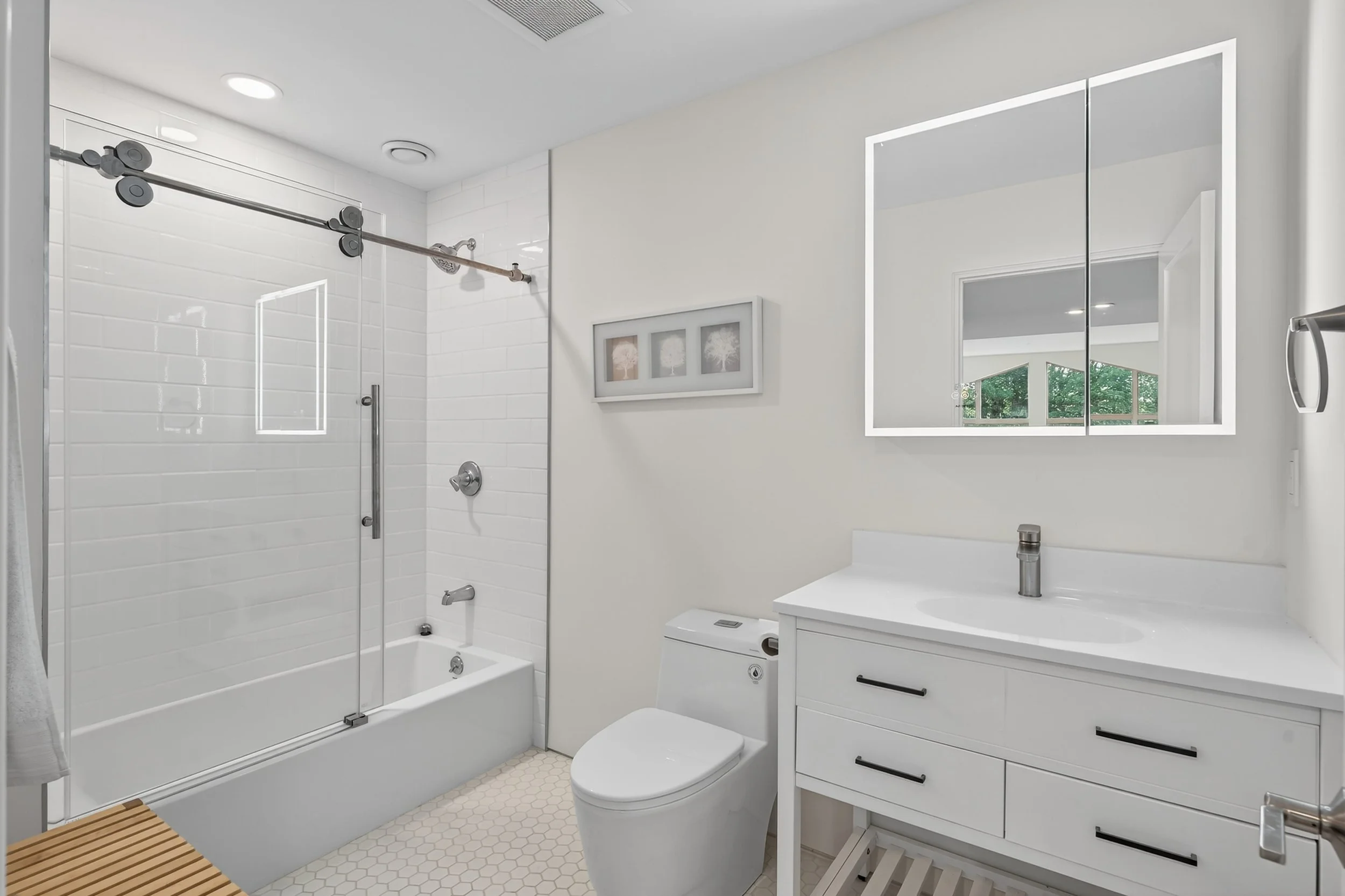 A modern bathroom with a glass-enclosed shower, a white vanity with a sink, a wall-mounted mirror with built-in lighting, and a small framed artwork on the wall. The room features white walls, a hexagon-patterned tile floor, and a window providing natural light.