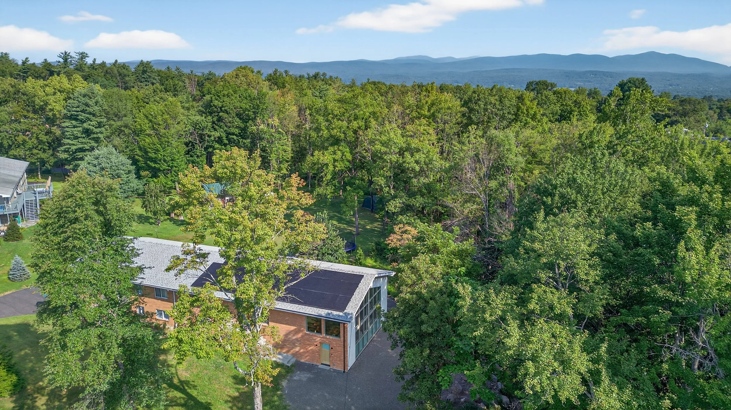 Aerial view of a residential neighborhood surrounded by dense green trees with mountains and blue sky in the background.