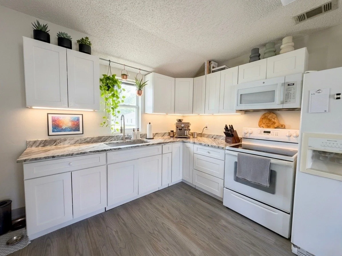 A modern kitchen with white cabinets, a marble countertop, a stainless steel sink, and a window with hanging plants. Appliances include a microwave, oven, and refrigerator. Decor includes potted plants and wall art.