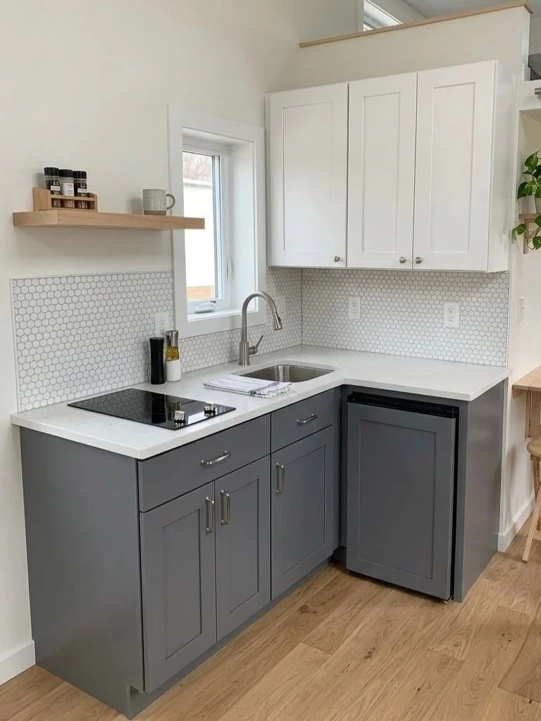 A small kitchen with gray lower cabinets, white upper cabinets, a white countertop, and a small sink. There is a window behind the sink, a black electric stovetop, a small mini fridge, a wooden shelf with spice jars, and a towel next to the sink.