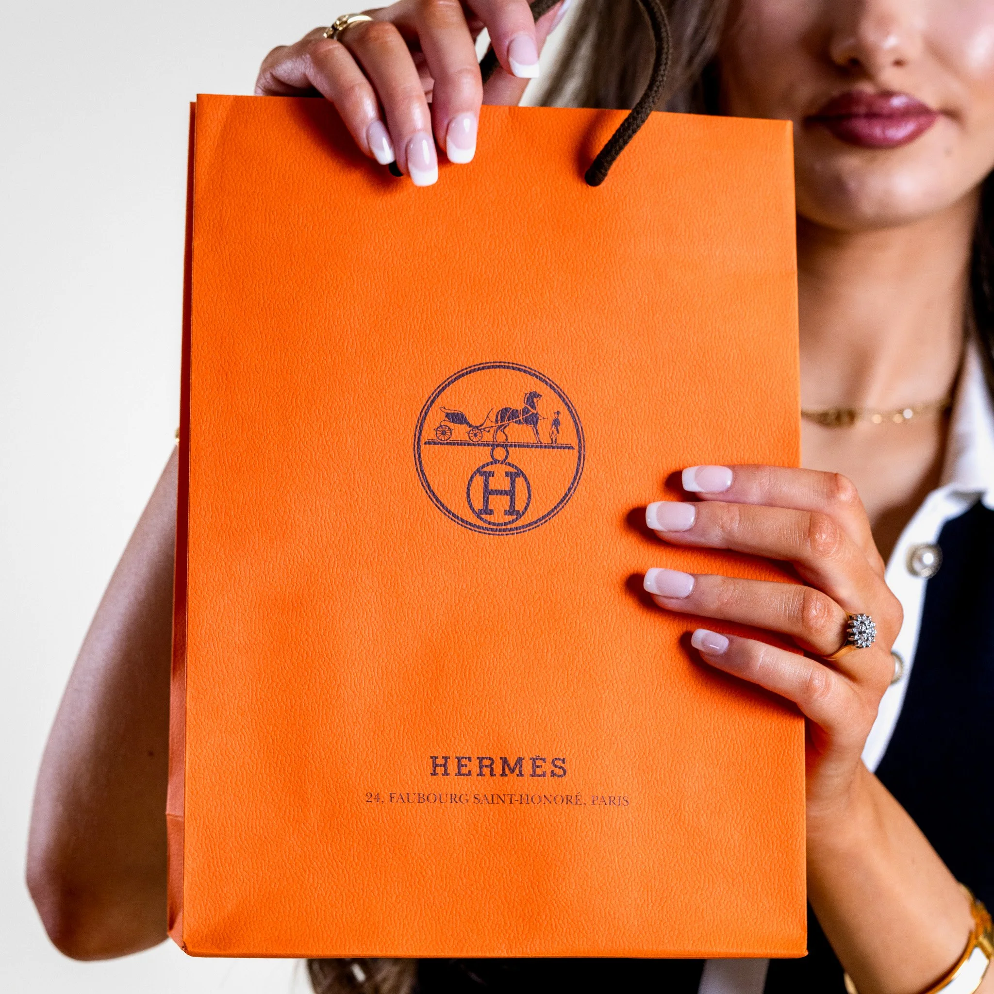 Person holding an orange Hermès shopping bag with logo and address, partially visible face, rings, and well-manicured nails.