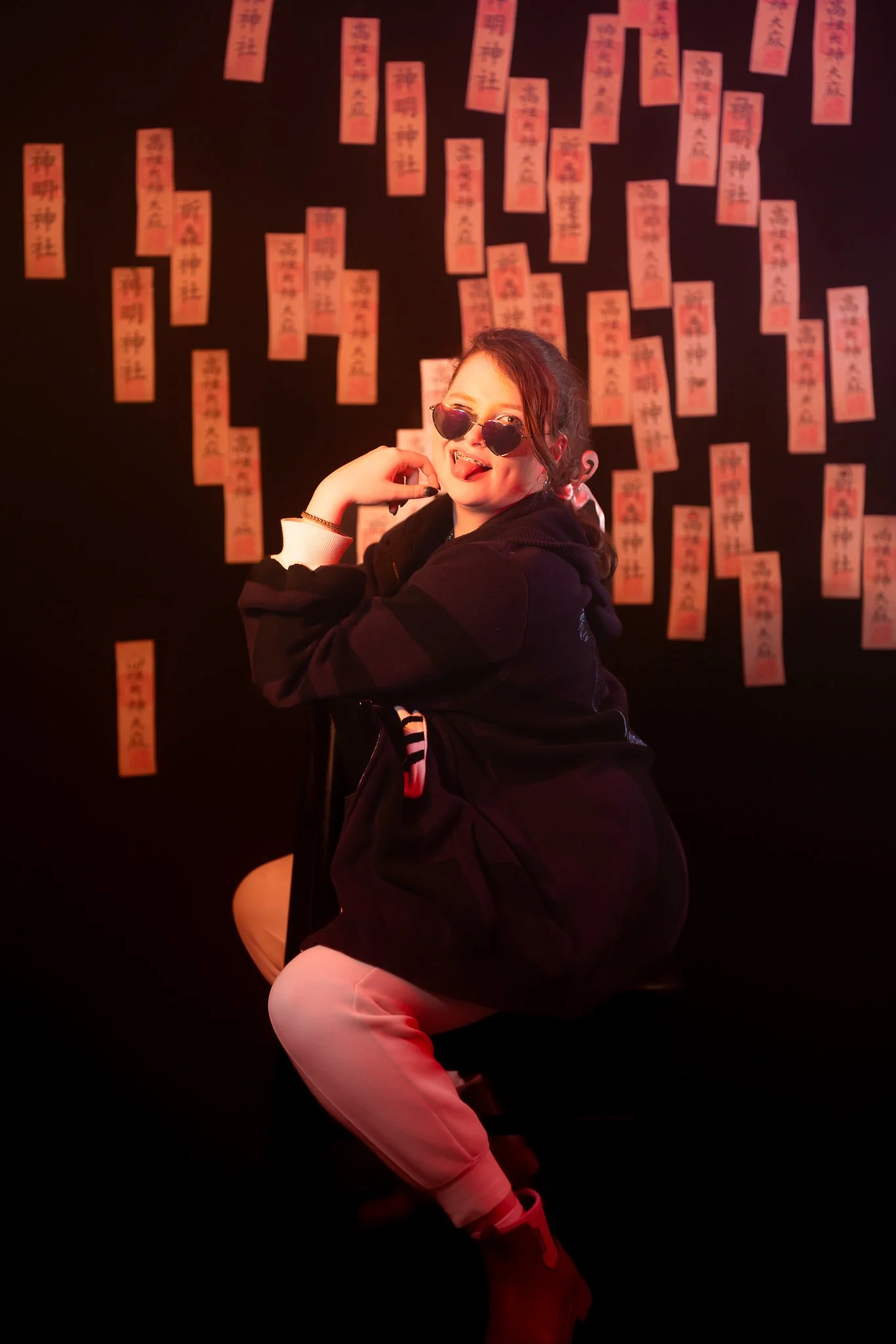 A young woman with sunglasses and a playful expression, wearing a black hoodie and beige pants, sitting on a chair against a dark background decorated with red paper strips with Asian characters.
