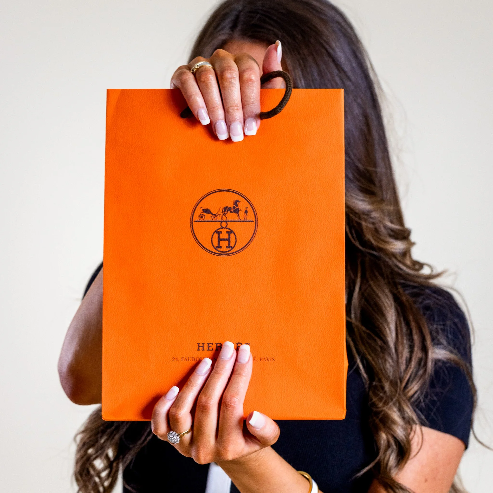 A woman with long wavy hair holding an orange Hermès shopping bag in front of her face, with a black background visible.