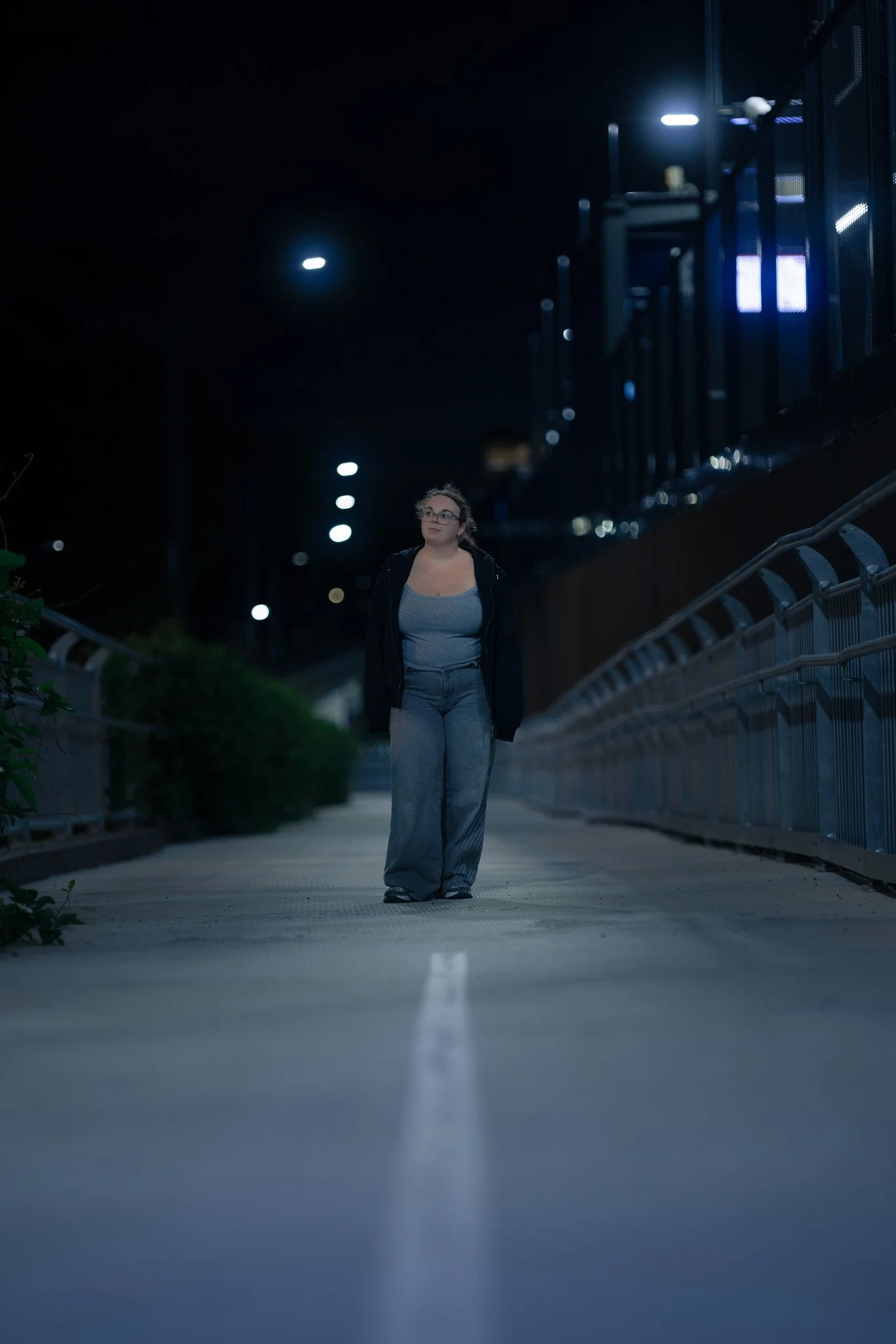 A woman walking alone on a dimly lit outdoor pathway at night, surrounded by a metal railing and illuminated streetlights in the background.