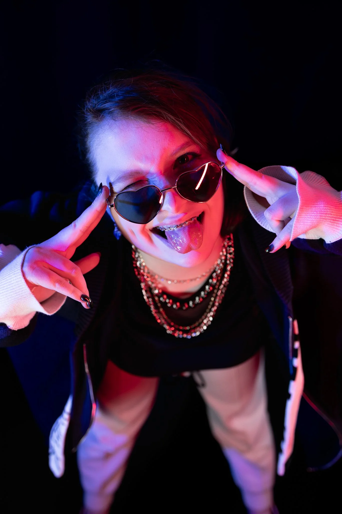 Person wearing heart-shaped sunglasses, sticking out tongue, making a peace sign with both hands, surrounded by colorful neon lighting, dressed in layered necklaces and a black jacket.