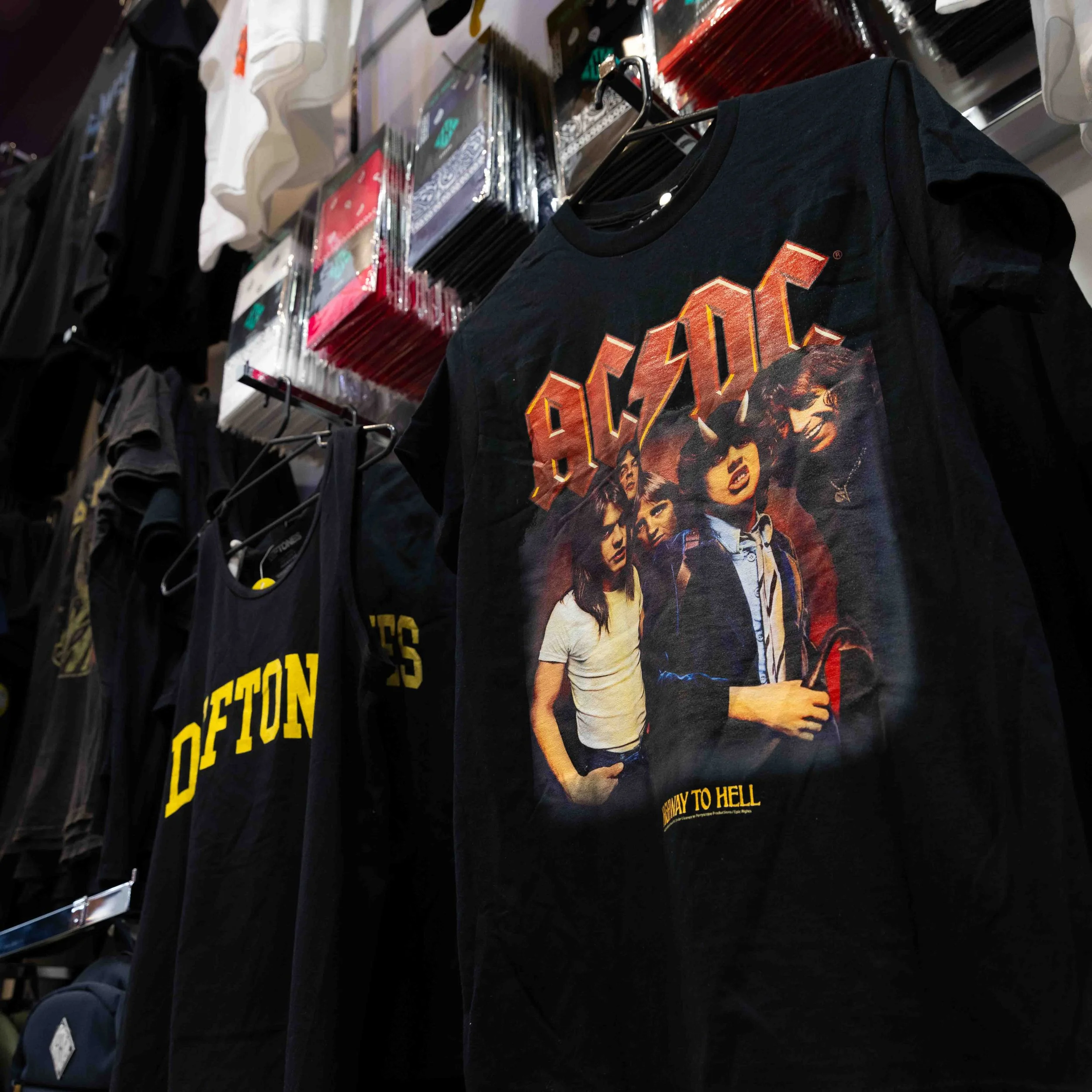 A black AC/DC band t-shirt featuring an image of the band members and the text 'Highway to Hell' hangs on a rack in a clothing store, with other band t-shirts and rectangular packages of socks displayed on shelves behind it.