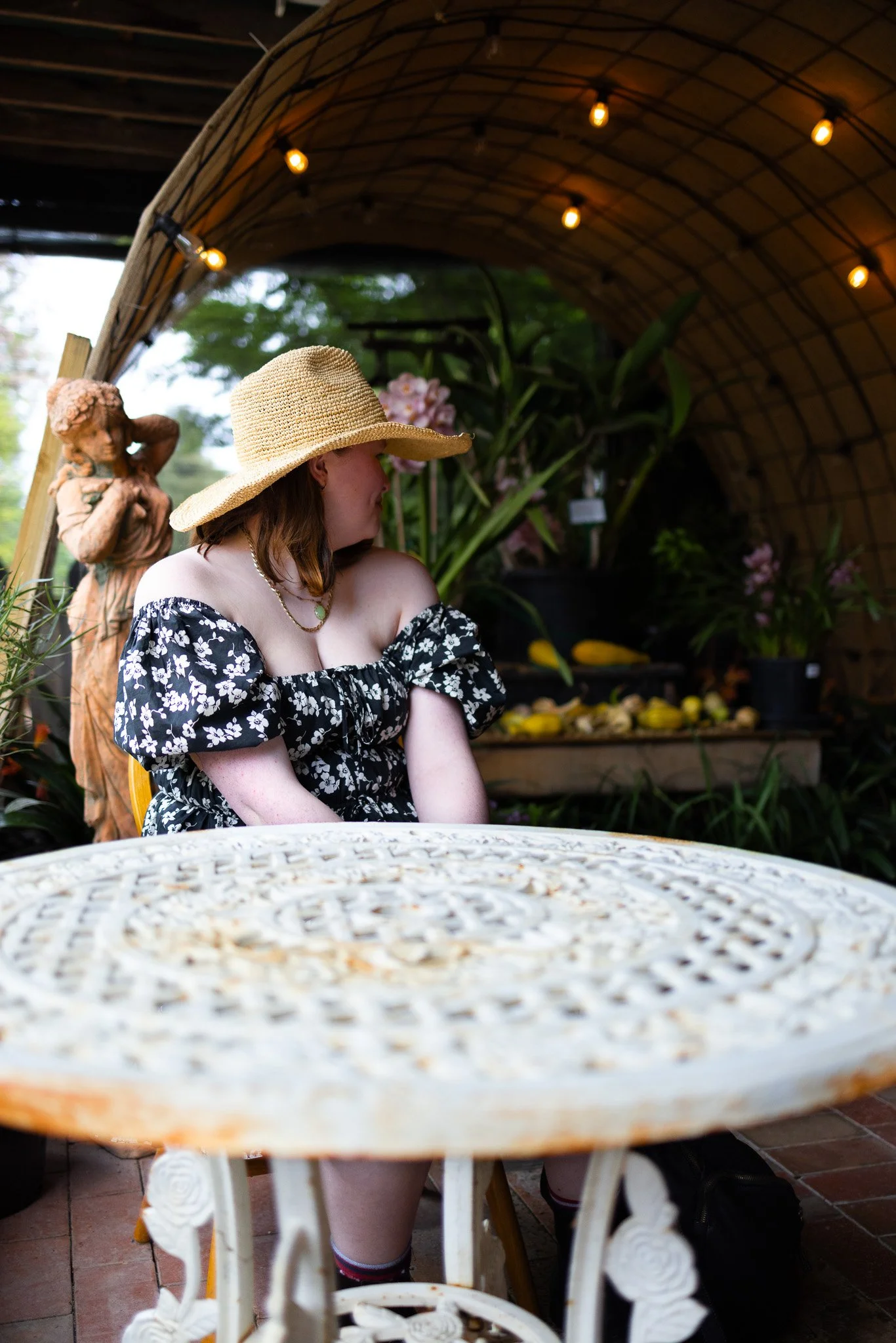 A woman in a black floral off-shoulder dress and a large straw hat sitting at a white ornate table in a garden with potted plants and a statue behind her, under a curved canopy with string lights.
