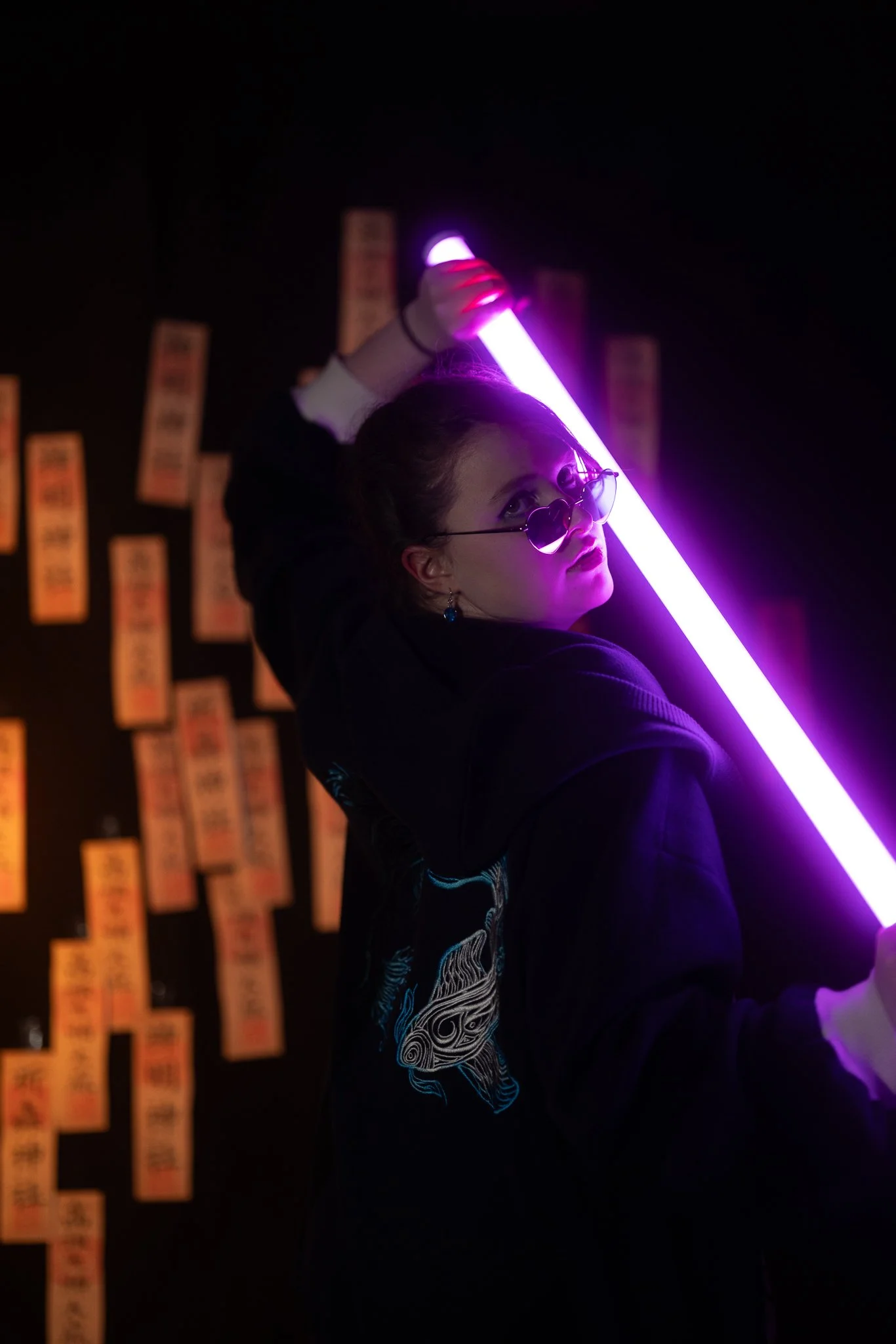 A young woman with glasses holding a glowing purple light stick in a dark room, with orange notes or tickets on the black wall behind her.