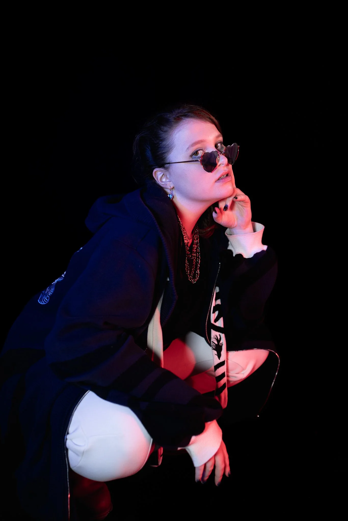 A woman squatting against a black background, wearing sunglasses, a dark hoodie, cream-colored pants, and layered necklaces, with a contemplative expression.