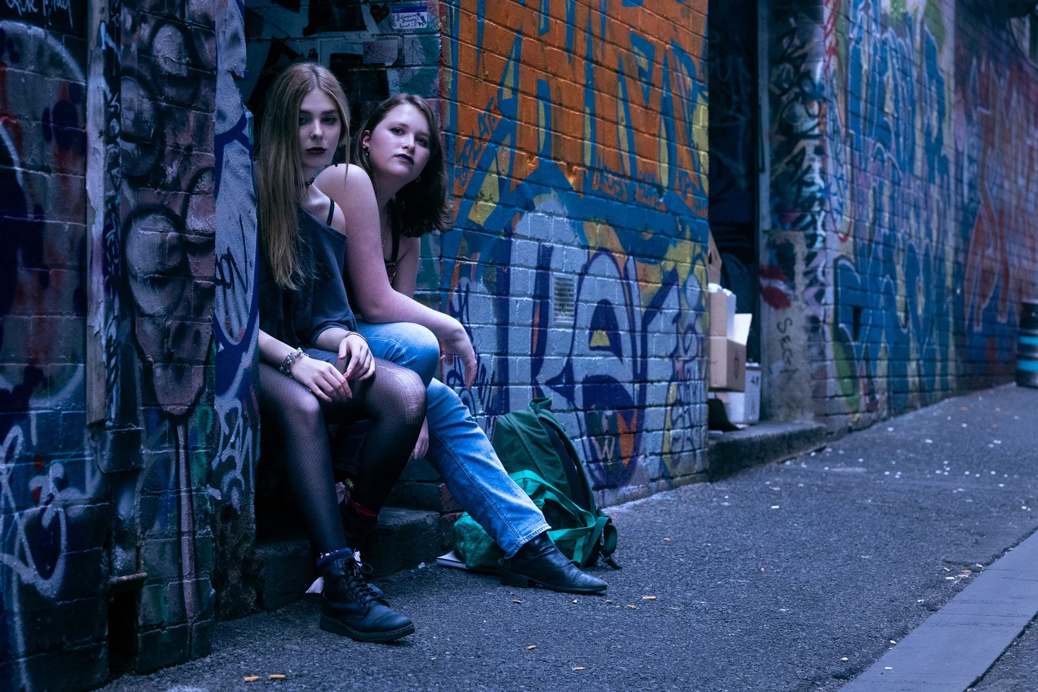 Two young women sitting on a curb against a graffiti-covered brick wall on a city street at twilight.