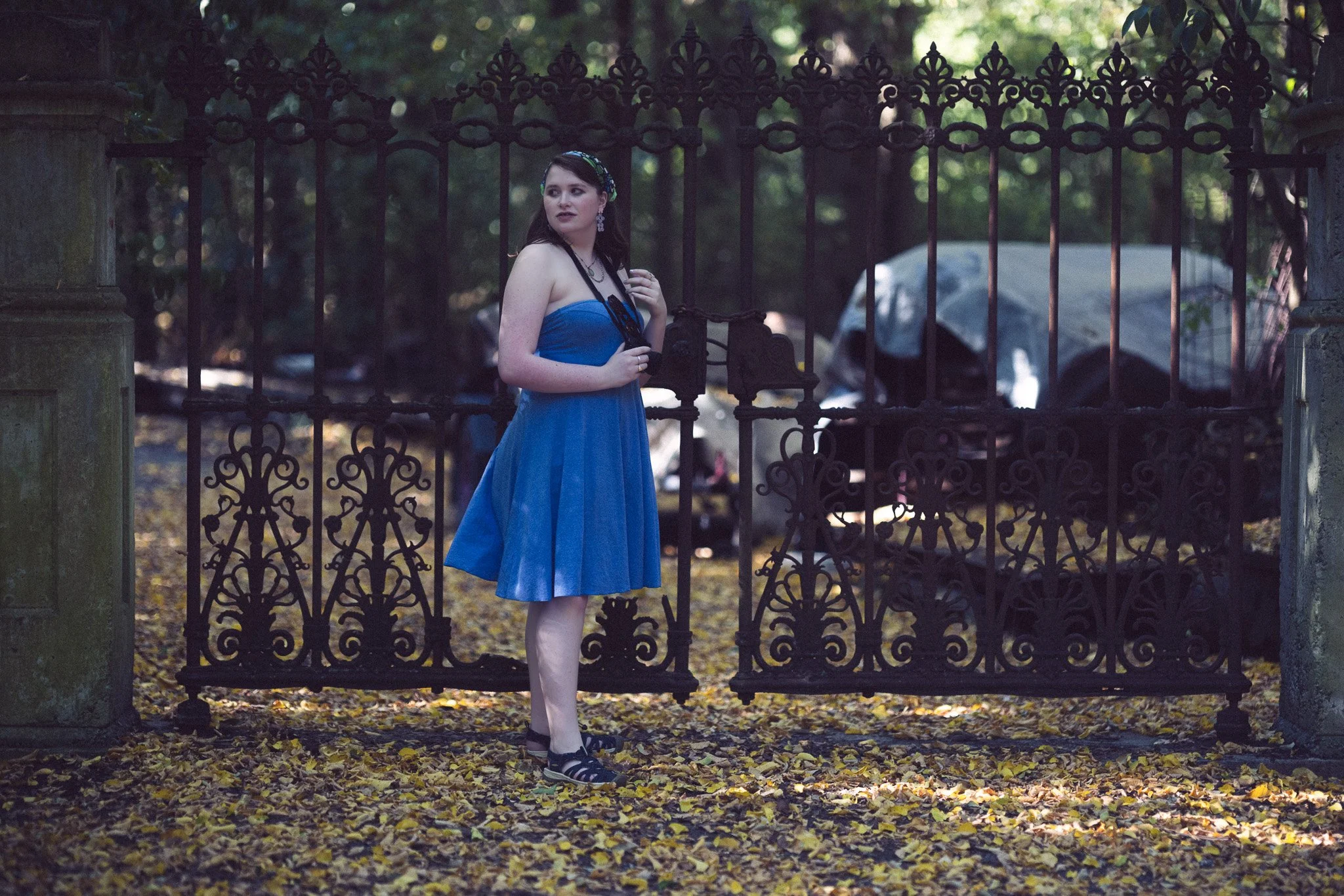 A young woman in a strapless blue dress stands behind an ornate black wrought iron fence, holding a black clutch and looking to her left, in a park with fallen yellow leaves and trees in the background.