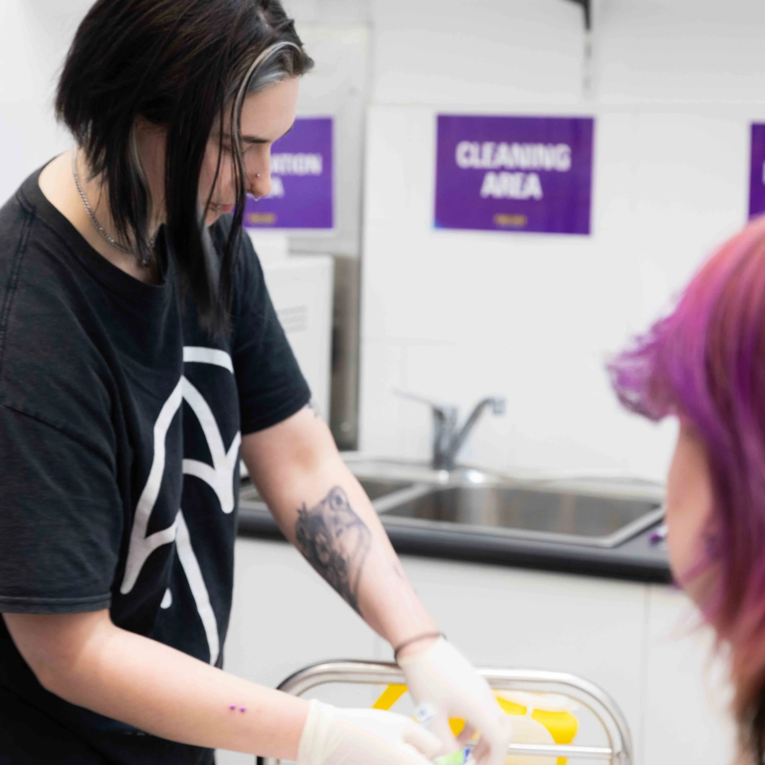 A woman with black hair and tattoos on her arms wearing a black T-shirt and white gloves, working at a station in a cleaning area, with a person with purple-dyed hair sitting in front of her. Signs in the background read 'CLEANING AREA'.