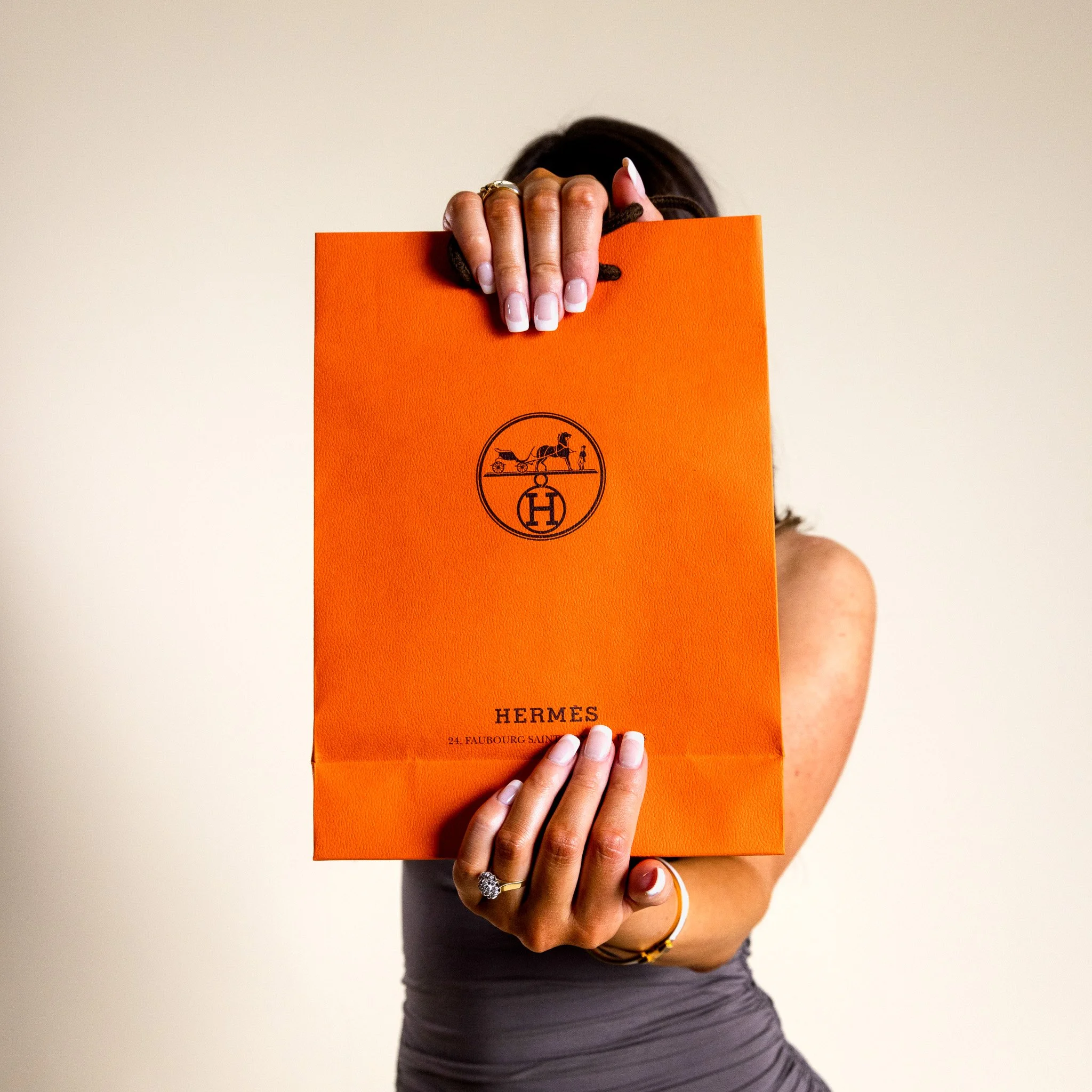 Person holding an orange Hermès shopping bag covering their face, with rifles and a horse logo printed on the bag, and the words 'Hermès' and an address at the bottom.