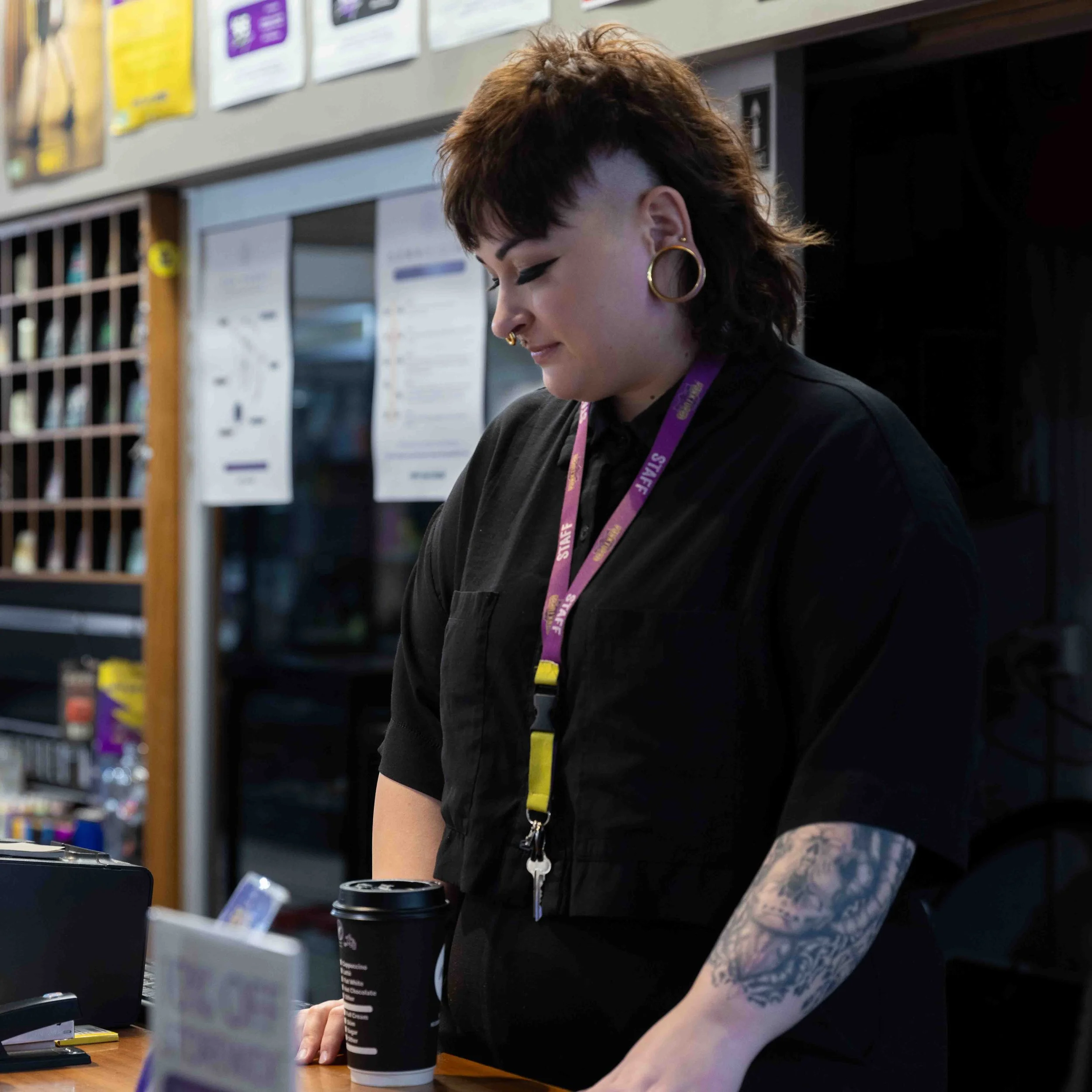 A woman with a mohawk hairstyle, multiple earrings, and tattoos on her arm, working at a counter in a coffee shop or restaurant.